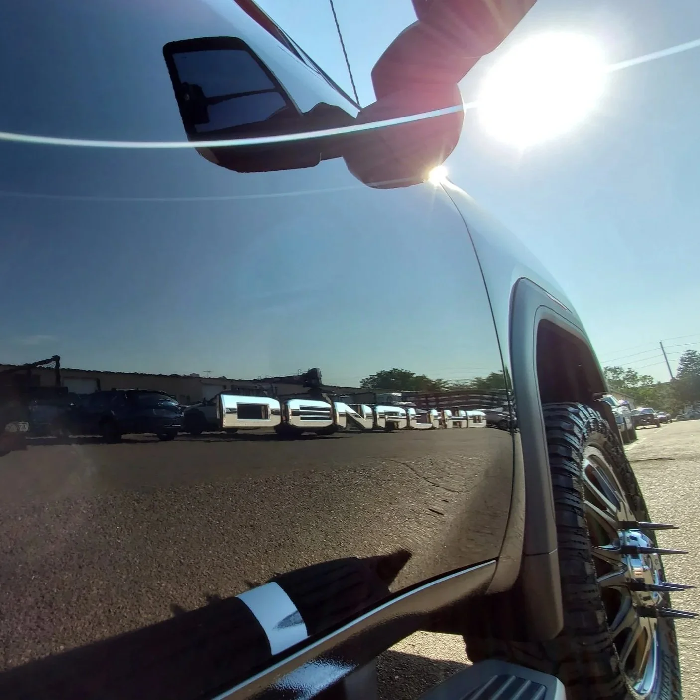 Close-up of a black vehicle's shiny exterior reflecting cars, people, and the sky, with a visible side mirror and wheel, taken outdoors on a sunny day.