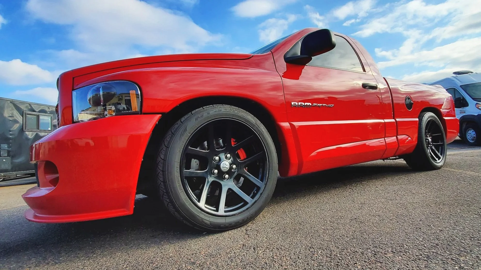 Red RAM SRT-10 pickup truck parked in a lot, with other vans in the background, under a blue sky with scattered clouds.