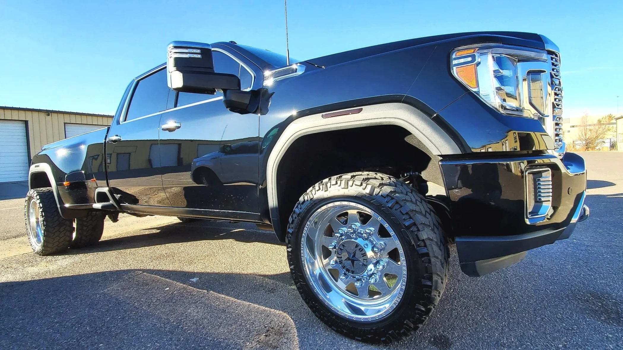 A black pickup truck with chrome wheels parked outdoors on a sunny day, reflecting the surrounding buildings and blue sky.