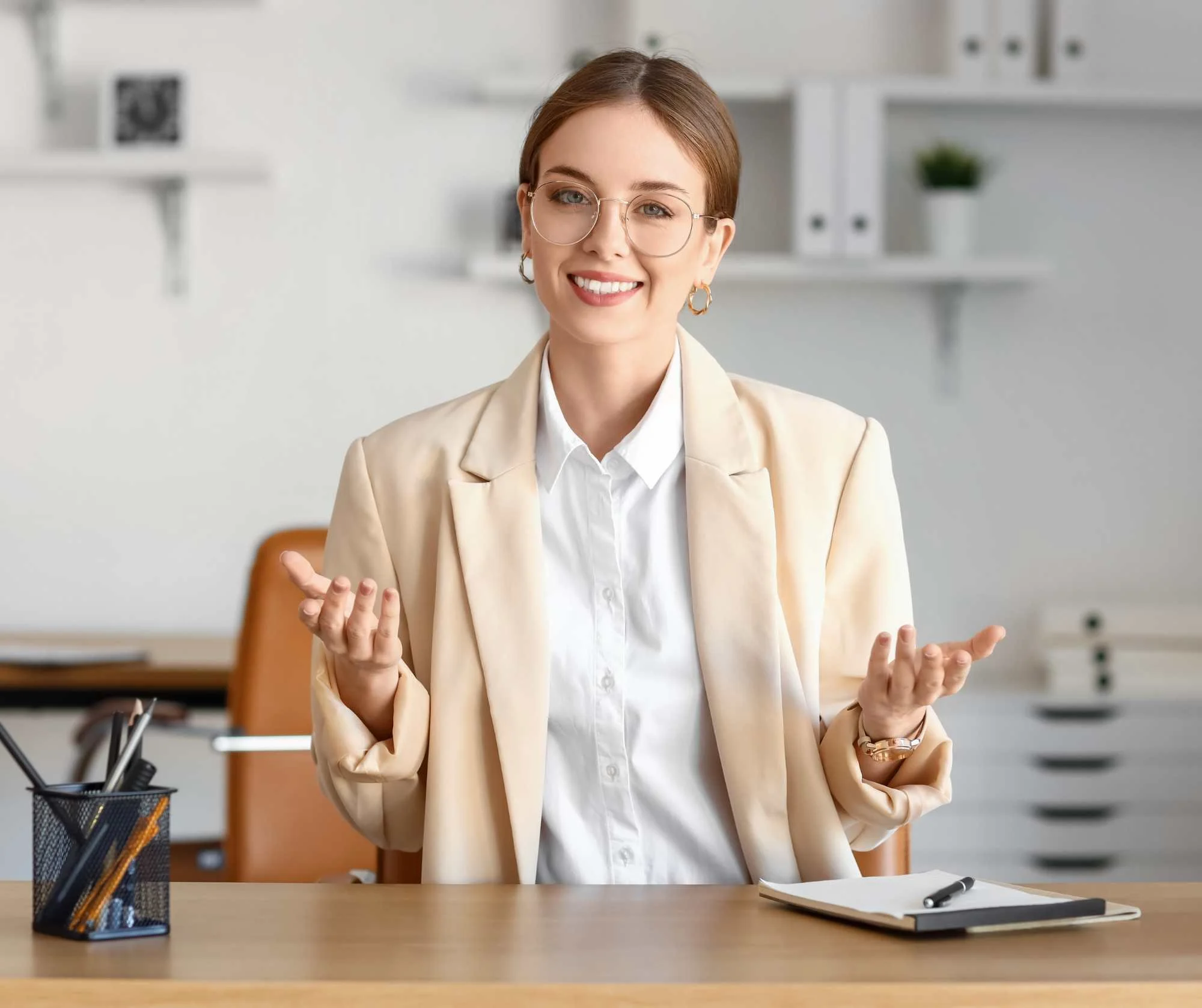 Smiling woman in a beige blazer speaking with open hands at a desk