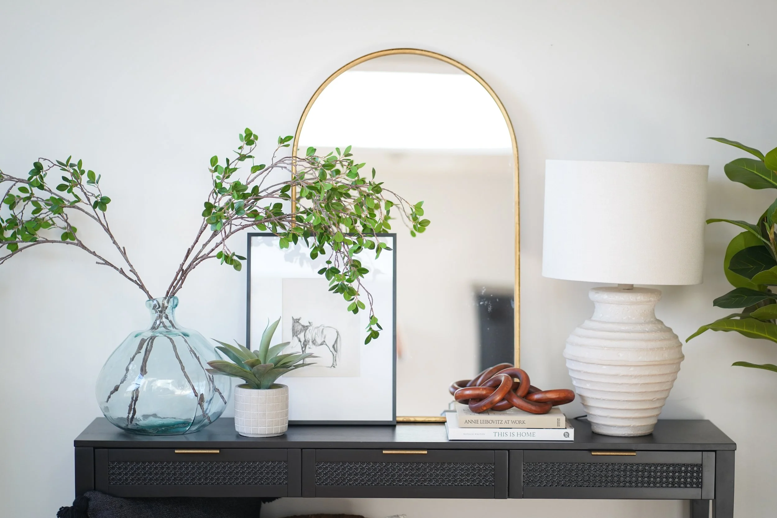 home staging Indianapolis, Indiana and short term rental styling and design. black console with cream lamp, large clear vase, gold mirror, wooden chain, books, framed drawing of a horse, home staging, interior design stager designer 