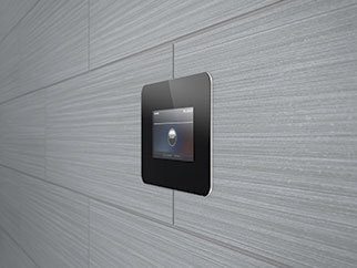 A modern black digital device mounted on a light gray tiled wall, displaying a circular pattern on its screen.