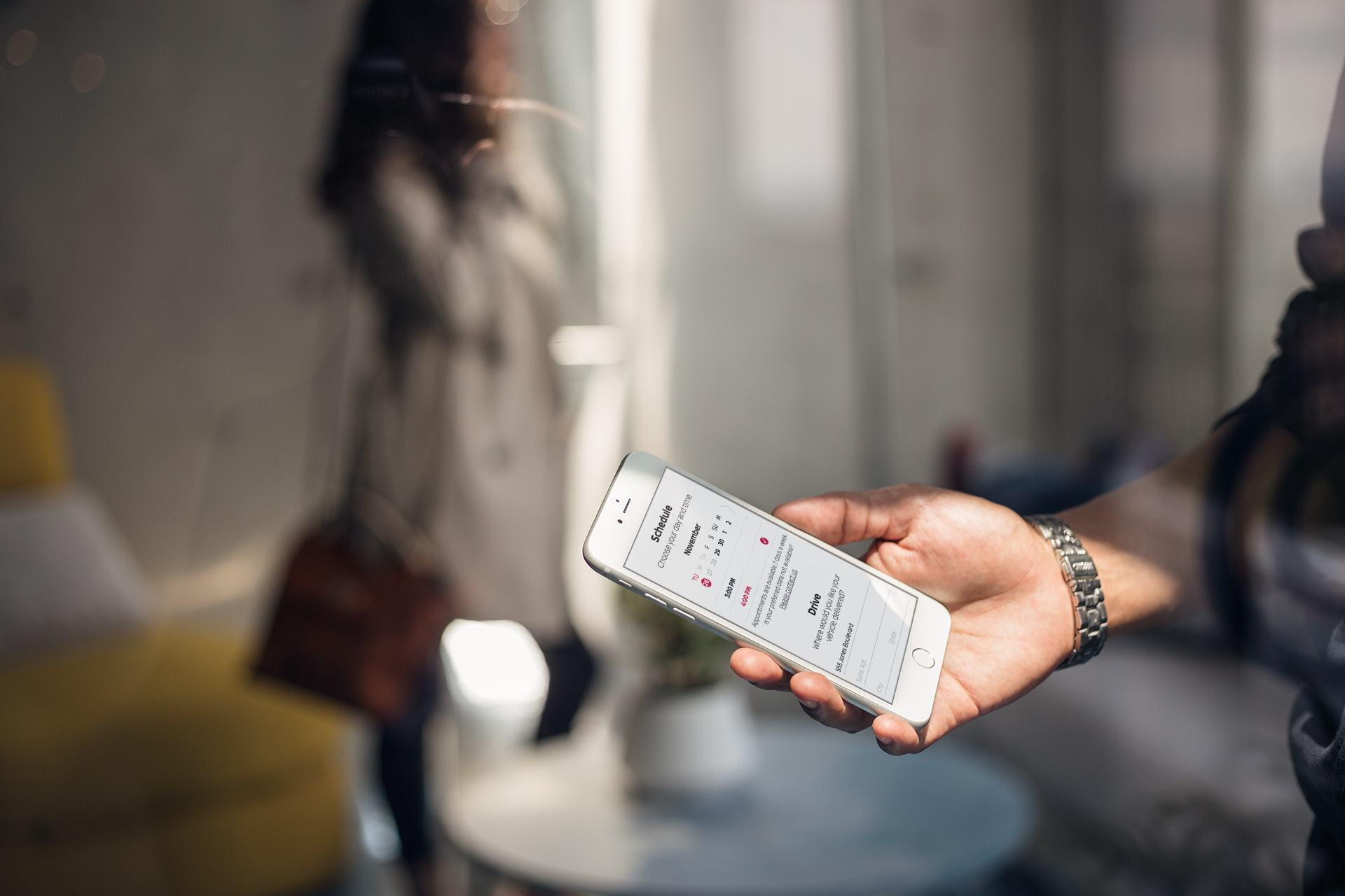Person holding a smartphone displaying a schedule or calendar app for Nissan Drive@Home service in a bright indoor setting.
