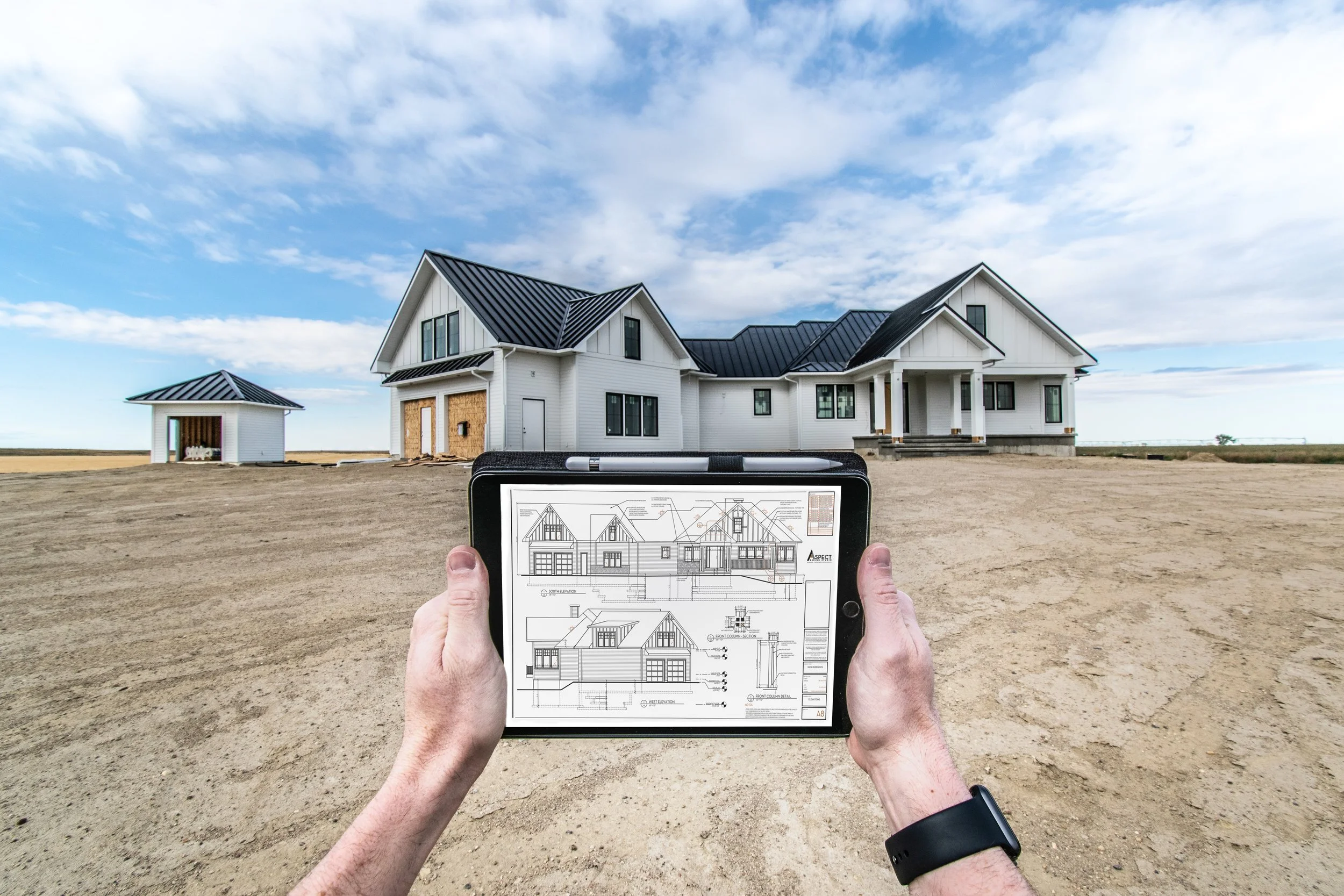 Person holding tablet displaying house blueprint in front of a new house under construction on a dirt lot.
