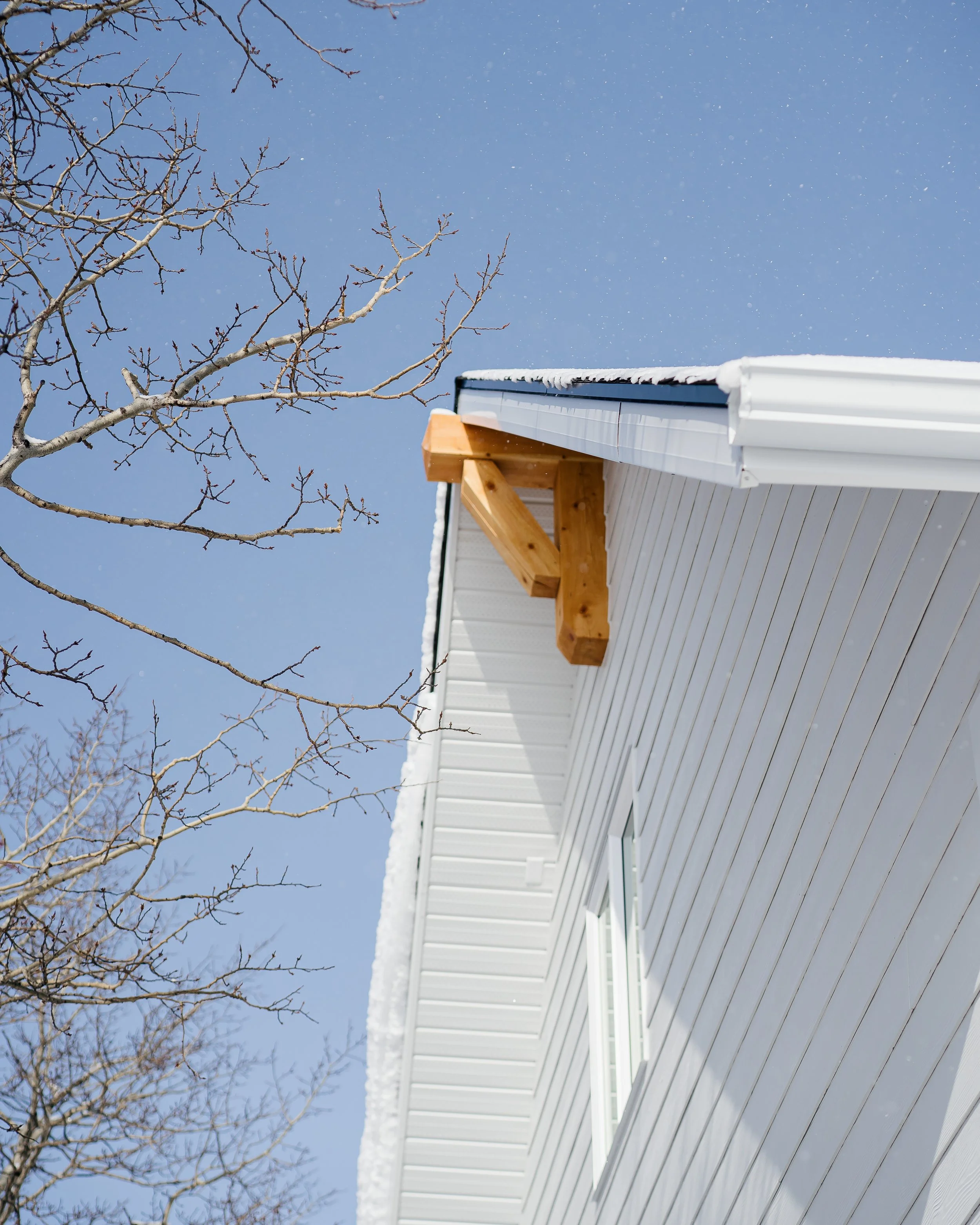 Close-up of the side of a white house with a window, snow on the roof, and an overhanging wooden beam, with a leafless tree and a clear blue sky in the background.