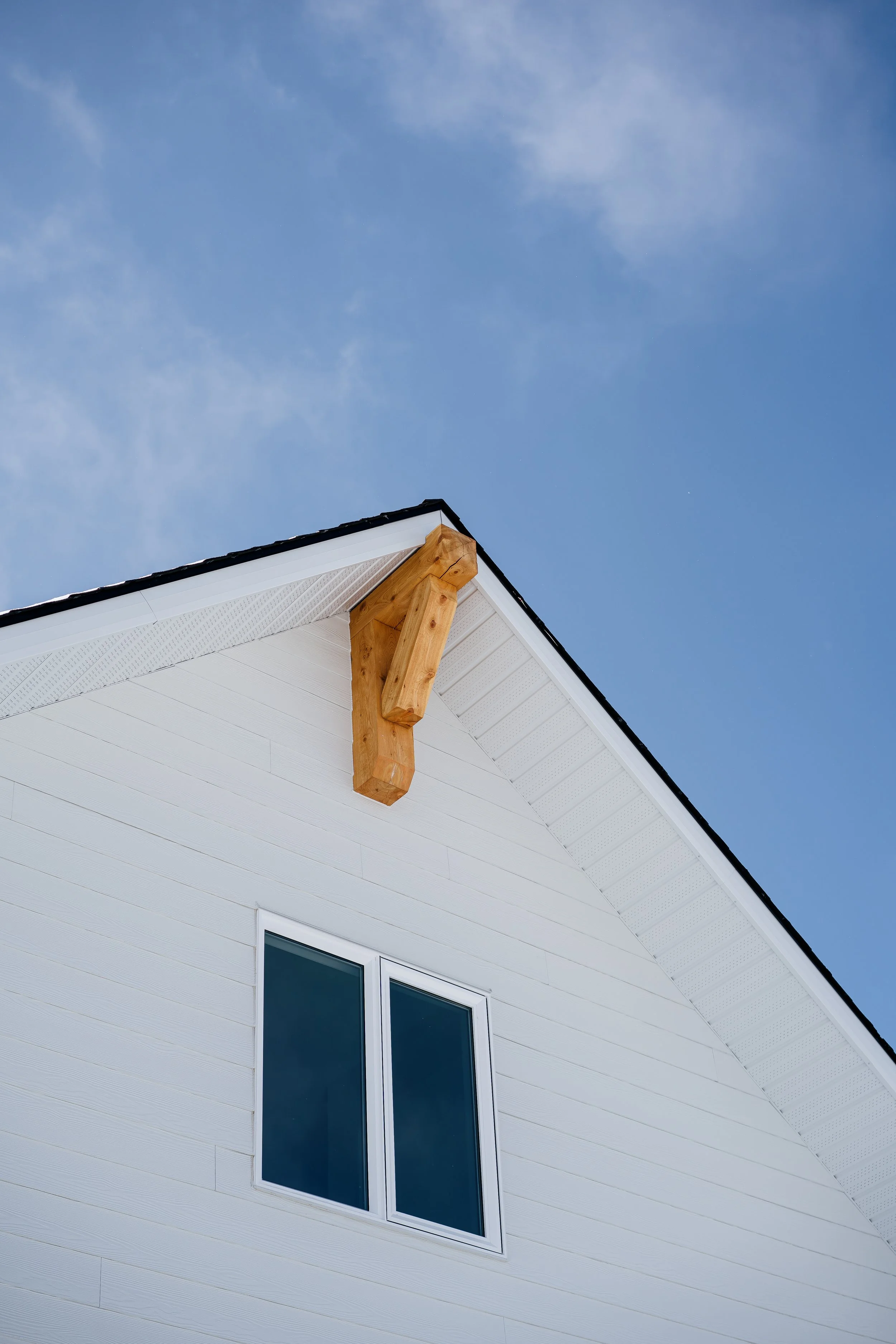 Close-up view of a white house's exterior corner with a window and a wooden support beam under the roof's overhang, against a clear blue sky.