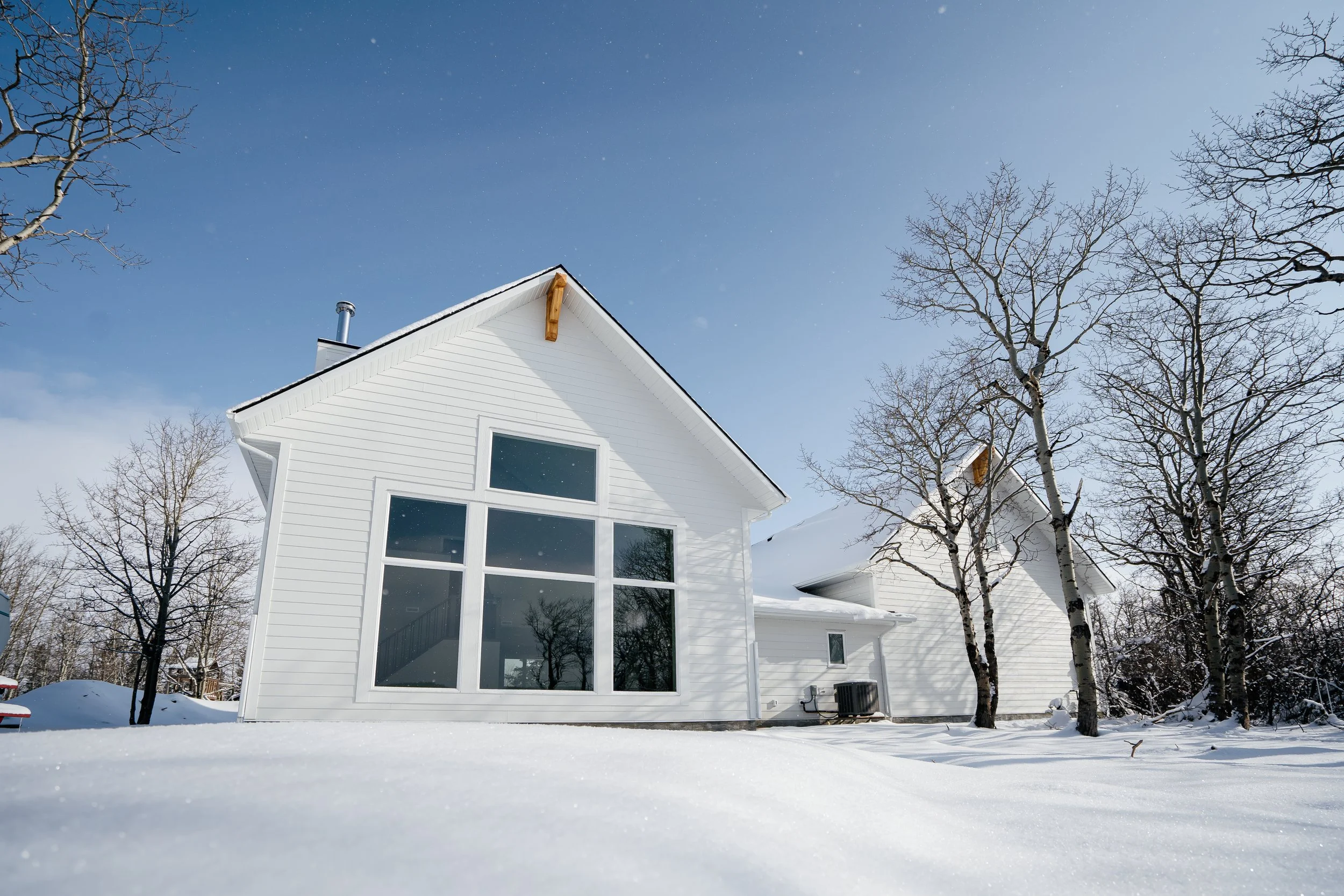 A modern white house with large windows and a steeply pitched roof, surrounded by snow and leafless trees under a blue sky.