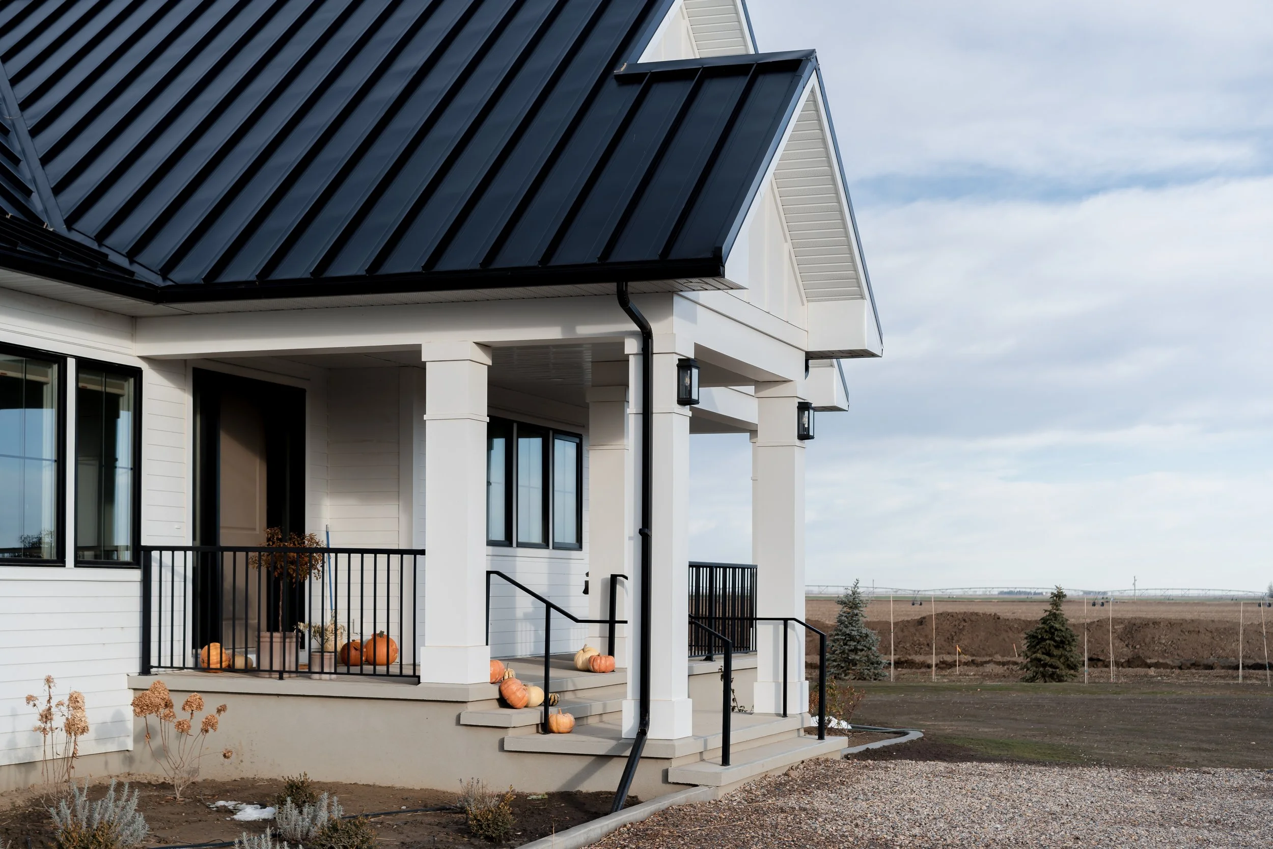 A white house with black metal roof, front porch decorated with pumpkins, and a rural landscape in the background.
