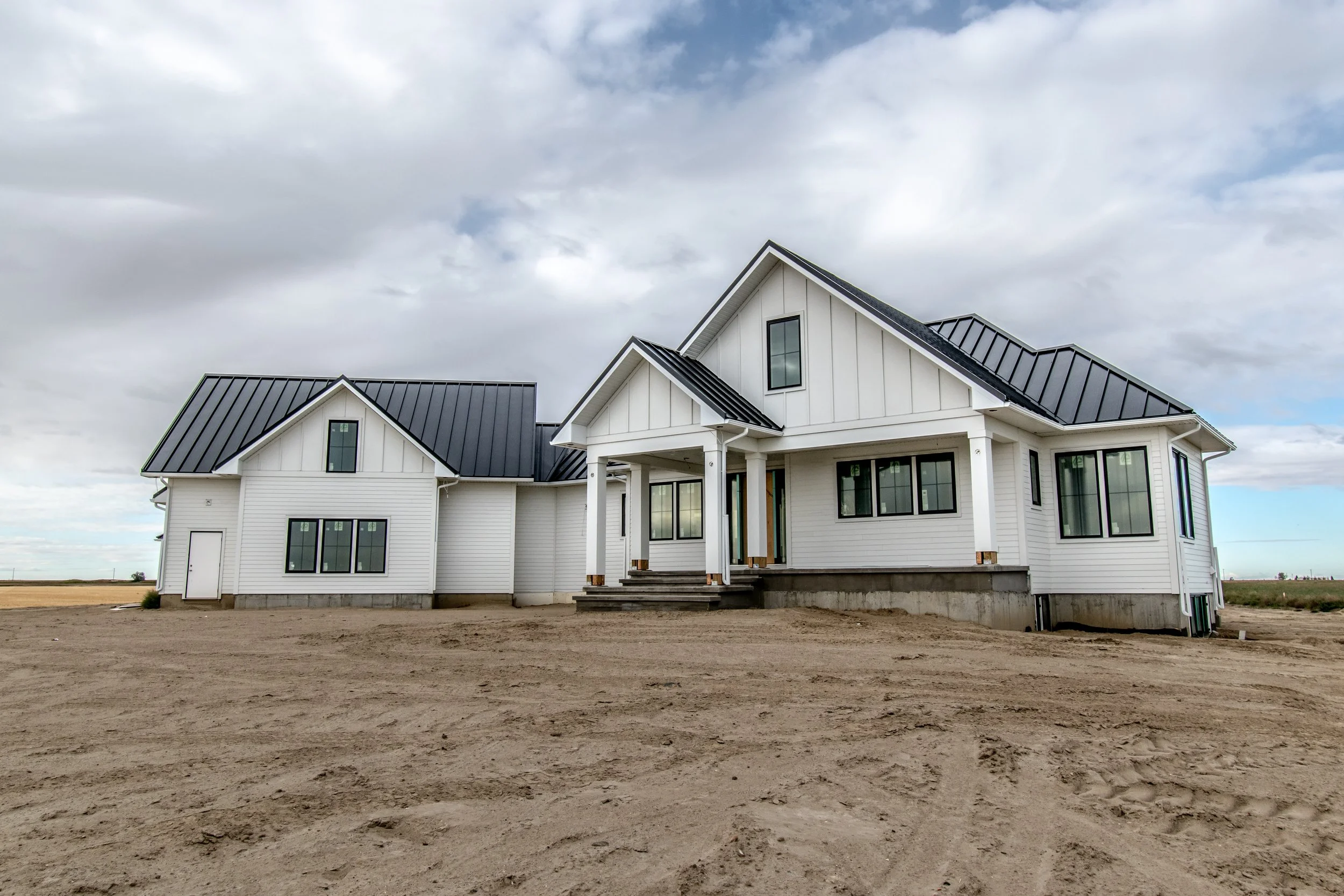 A modern white house with a black metal roof on a dirt lot under a cloudy sky.