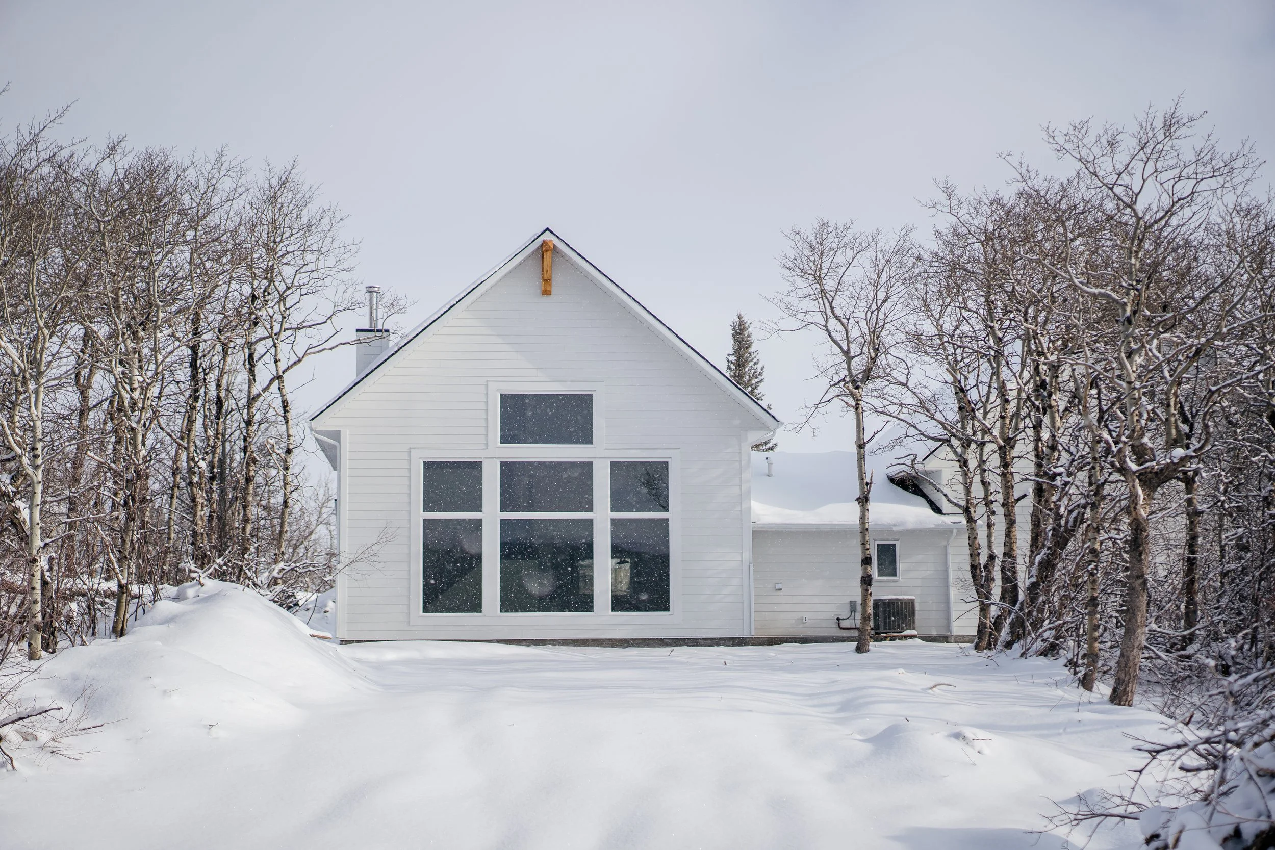 A white house with large windows on a snowy landscape, surrounded by leafless trees, during winter.