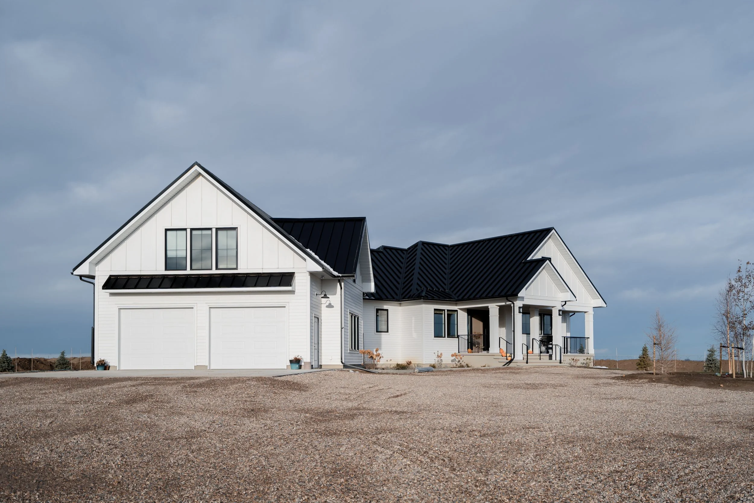 A modern white house with black metal roofing, two garage doors, and a front porch with steps, situated on a gravel lot under a cloudy sky.