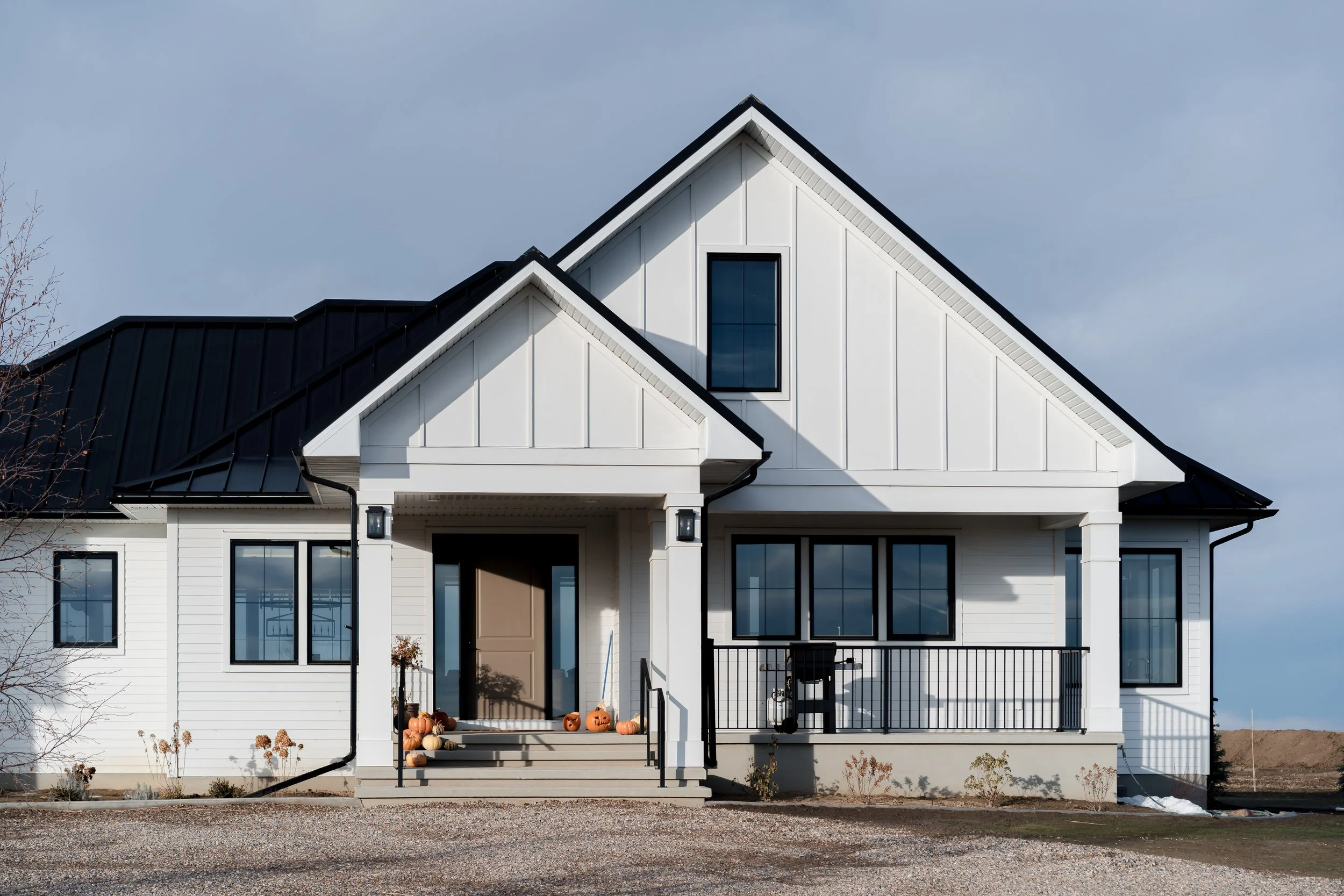 A modern white house with black roof, porch, and several pumpkins on the steps, indicating fall or Halloween season.