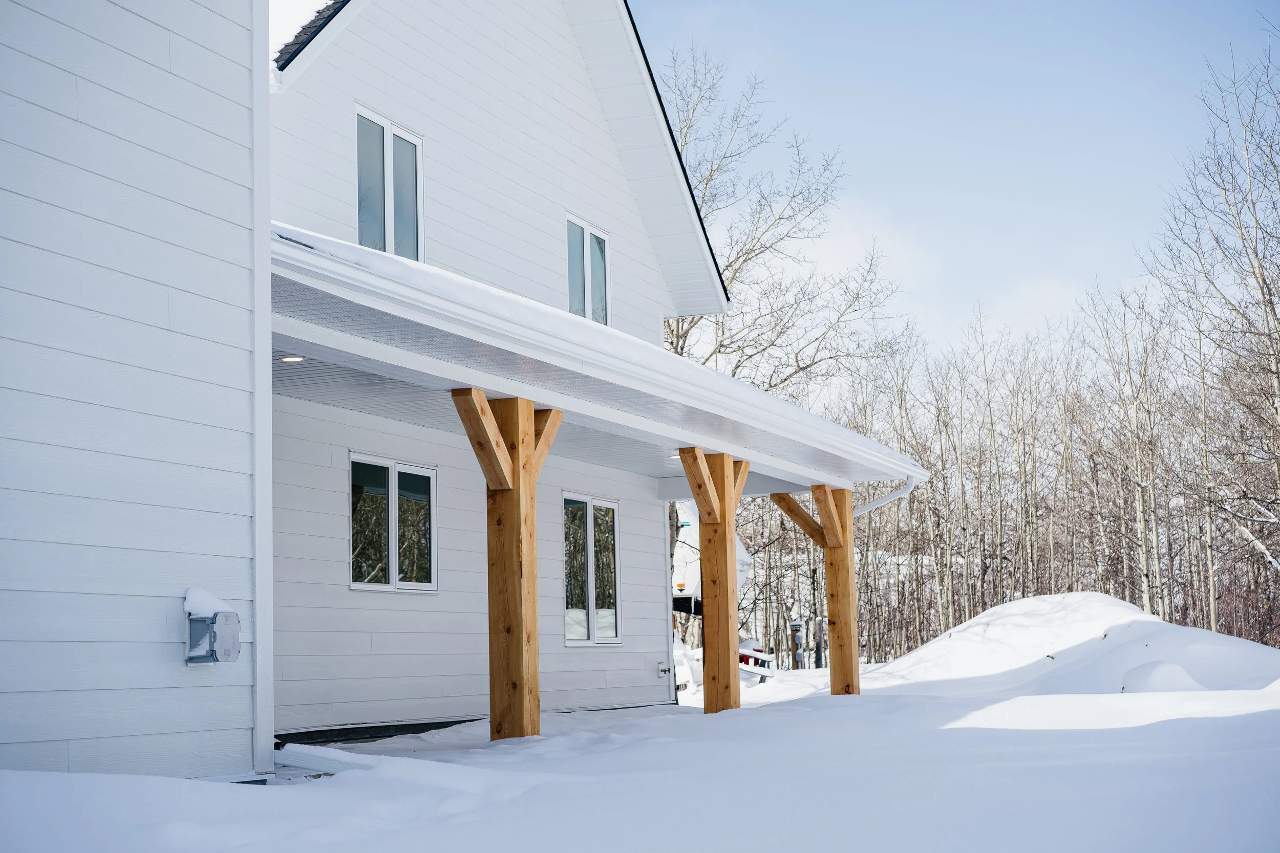 White house with wooden support beams under snow-covered porch, surrounded by snow and leafless trees, on a clear winter day.