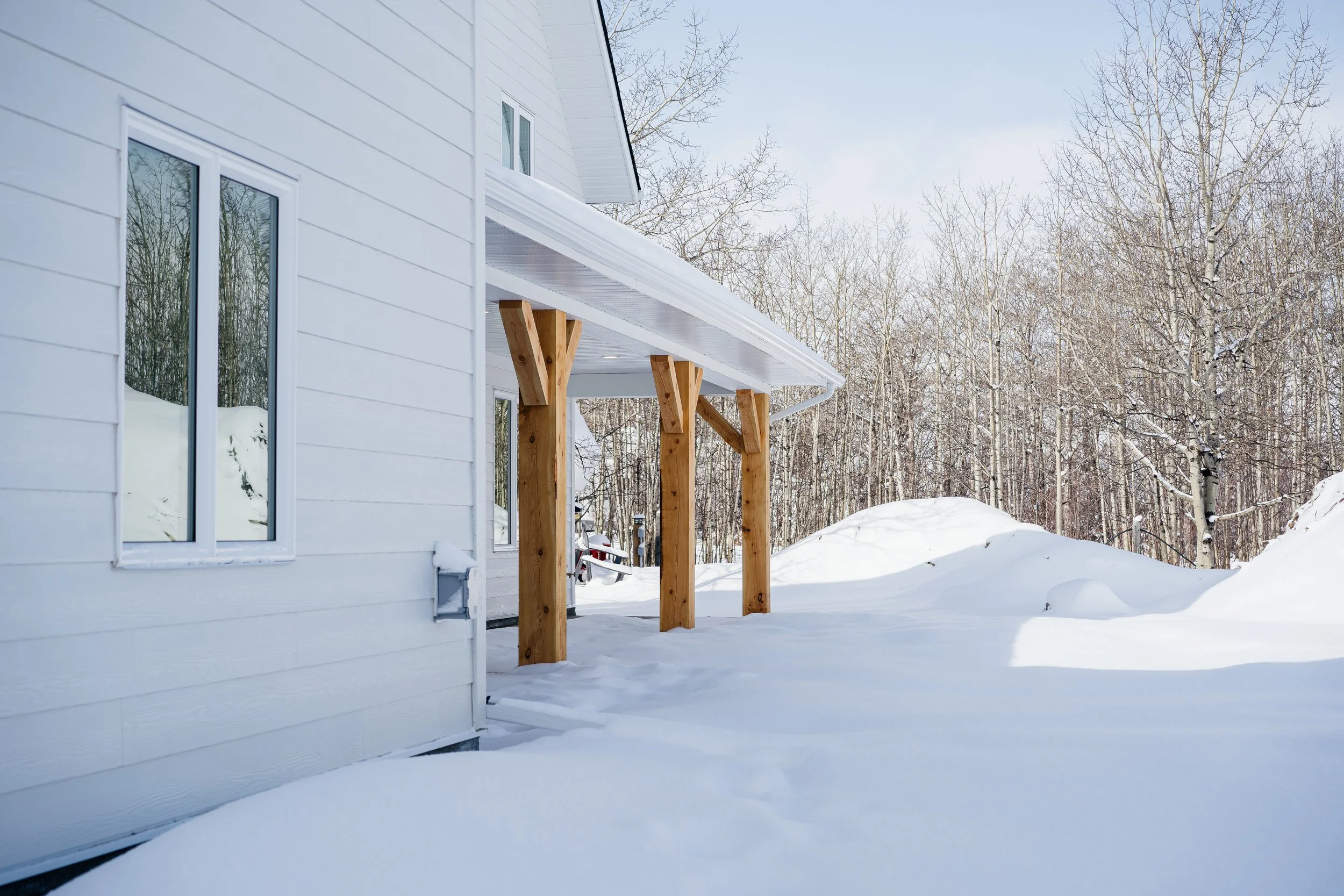 Snow-covered backyard with a white house featuring large windows, wooden support beams under a porch, and leafless trees in the background.