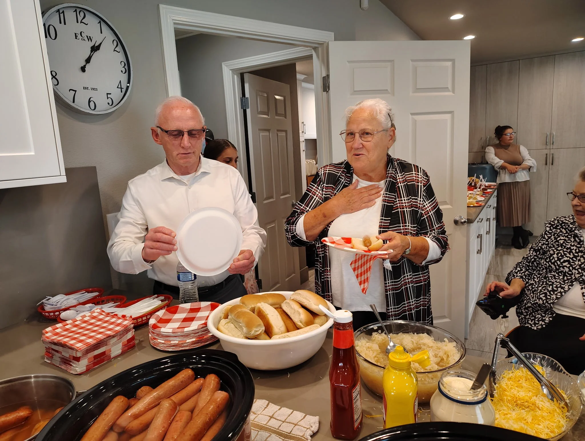 People at a buffet-style gathering serving hot dogs, sides, and condiments in a kitchen setting.