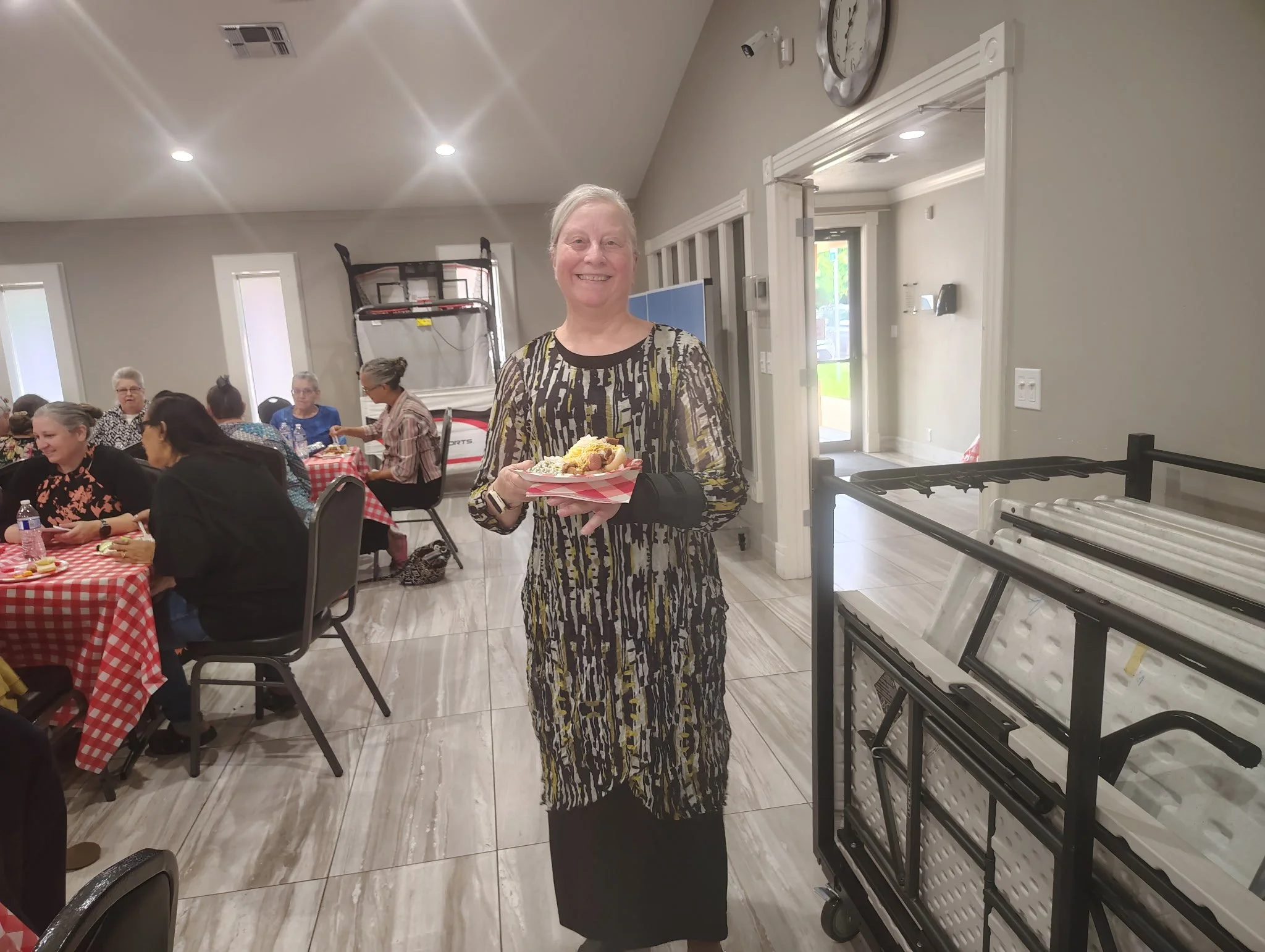 A smiling woman in a patterned dress holds a plate of food while standing in a room with people sitting at tables covered with red and white checkered tablecloths. There are windows, an exercise machine, and a cart nearby.