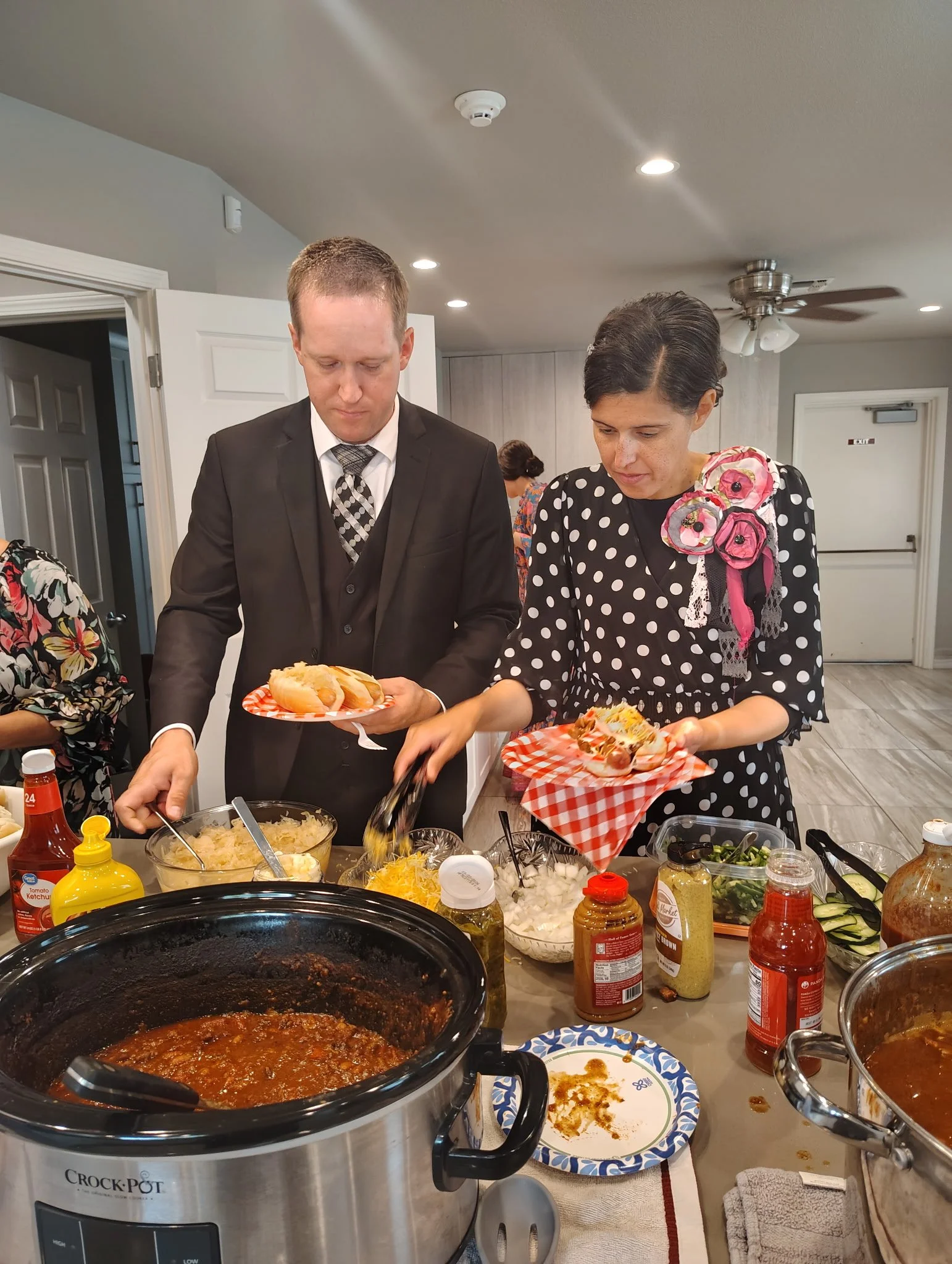 A man in a black suit and a woman in a black dress with white polka dots serving food at a buffet table with slow cooker chili, condiments, salad ingredients, and hot dogs.