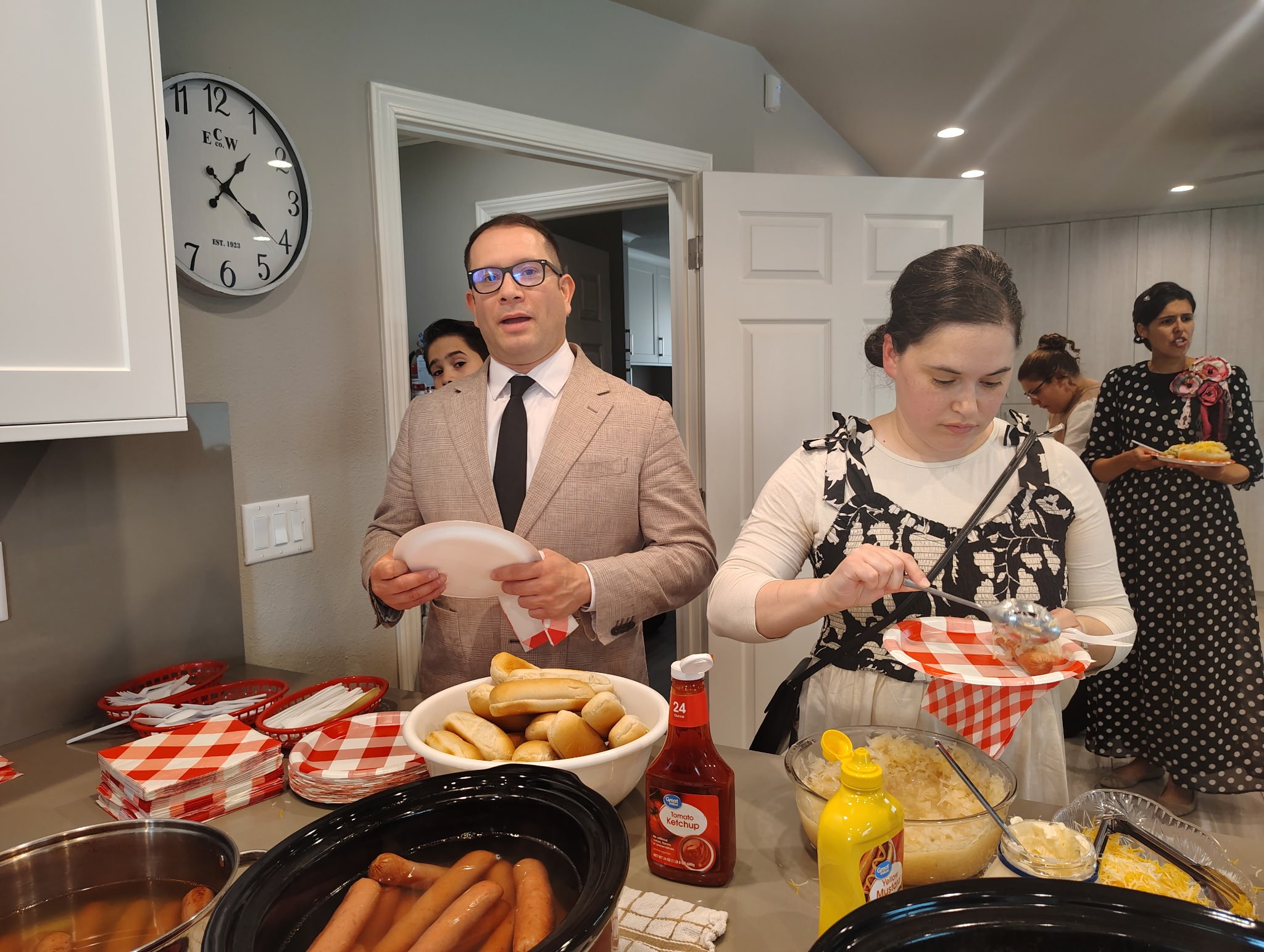 People preparing and serving food at a buffet table in a home kitchen, including hot dogs, buns, ketchup, mustard, and various side dishes.