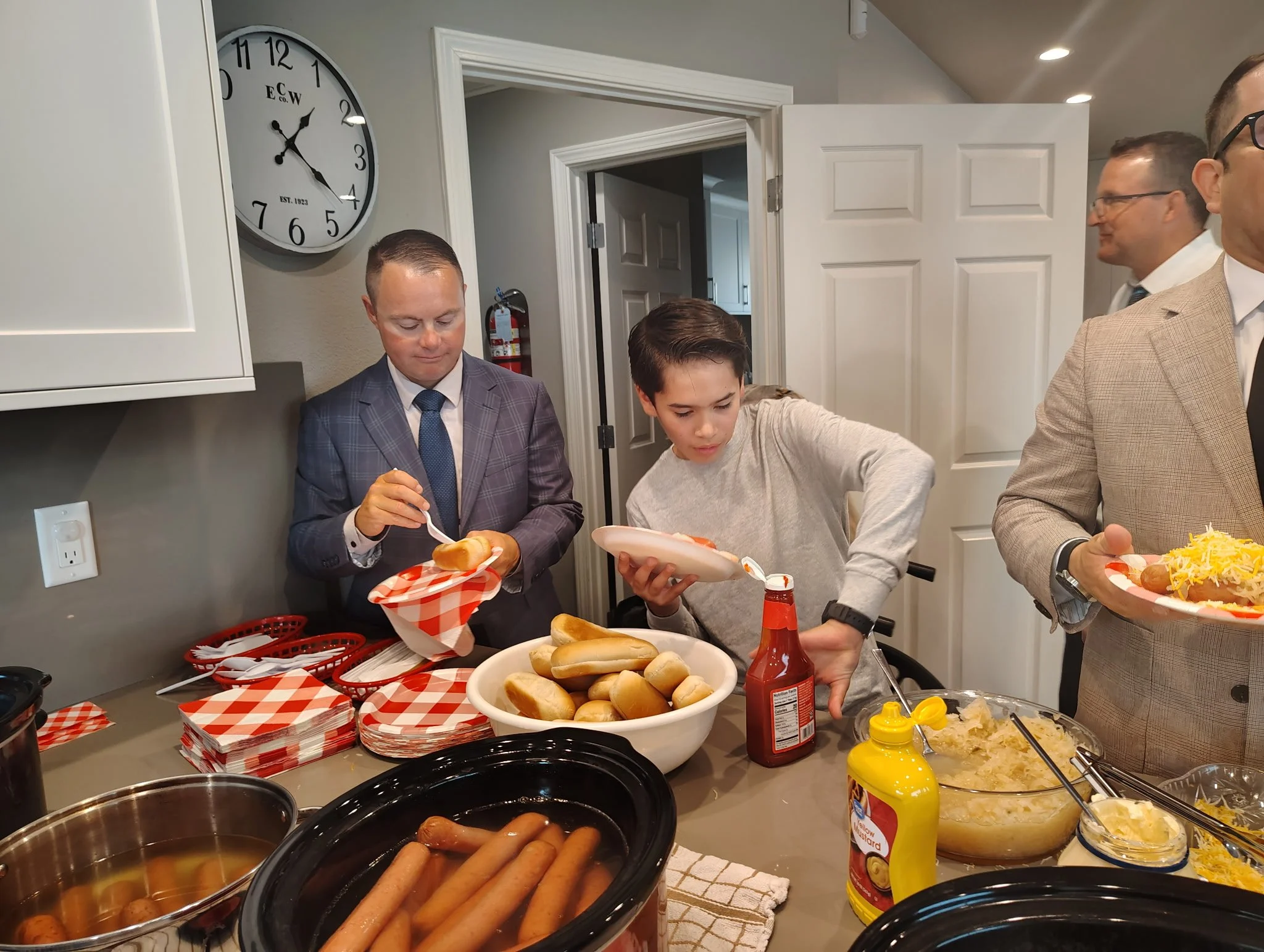 People serving and preparing hot dogs and food at a buffet table in a kitchen.