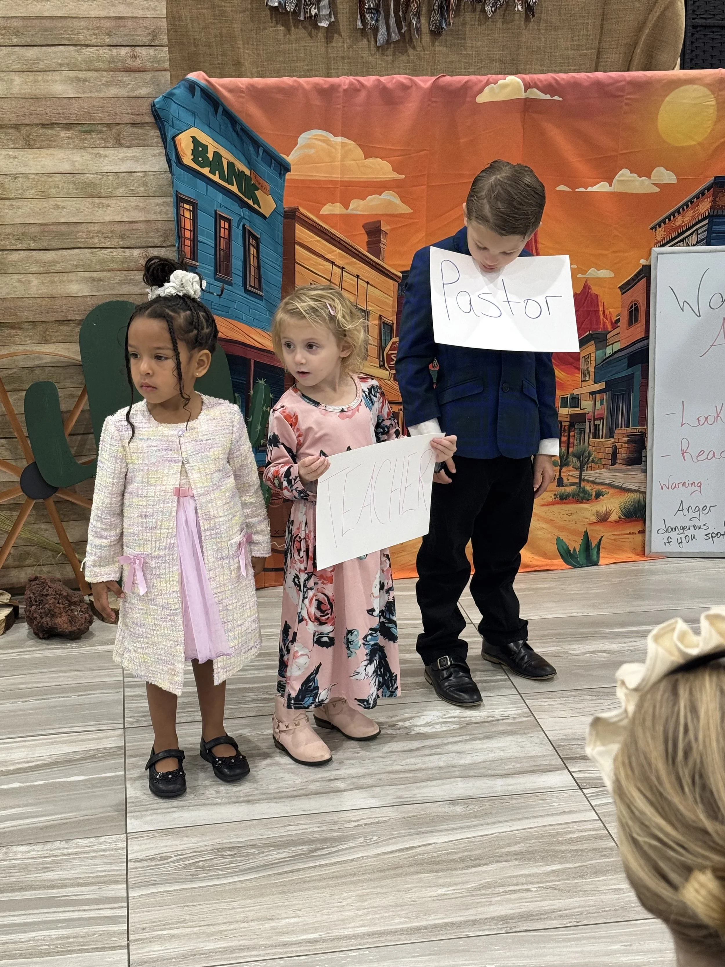 Three children standing on a stage holding signs with words, with a western-themed background and a wooden wall. The signs read 'Teacher', 'Pastor', and the third child's sign is not visible.