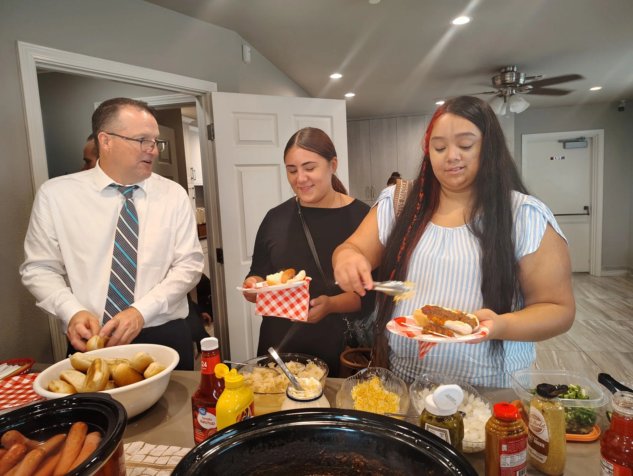 A group of people serving themselves food at a buffet table with hot dogs, buns, condiments, and toppings at an indoor gathering.
