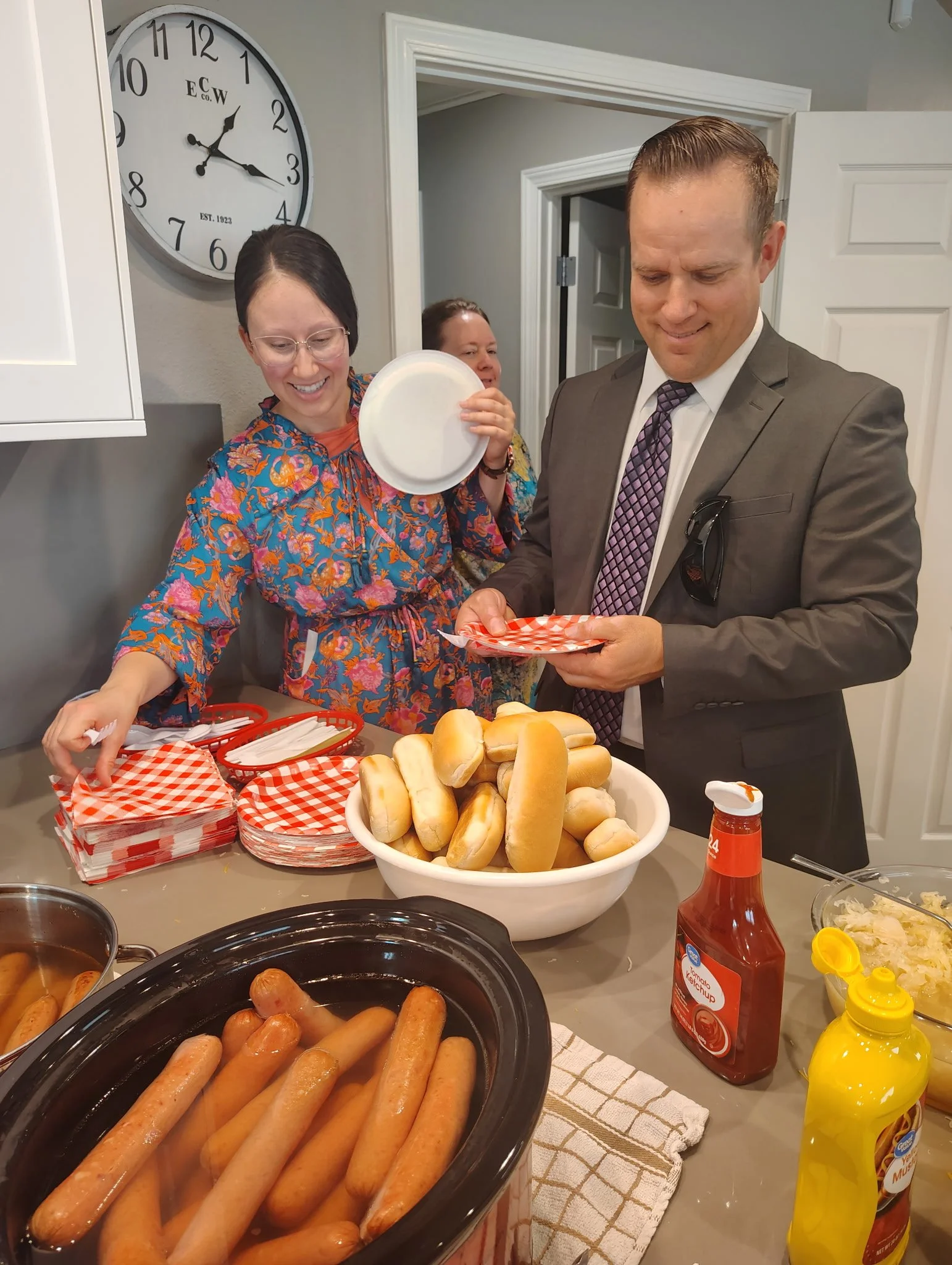 People gather around a table at a cookout, with hot dogs, buns, and condiments visible. A man in a suit and a woman in glasses and a colorful dress are smiling, holding paper plates and waiting to serve themselves. A large clock shows the time as 2:1