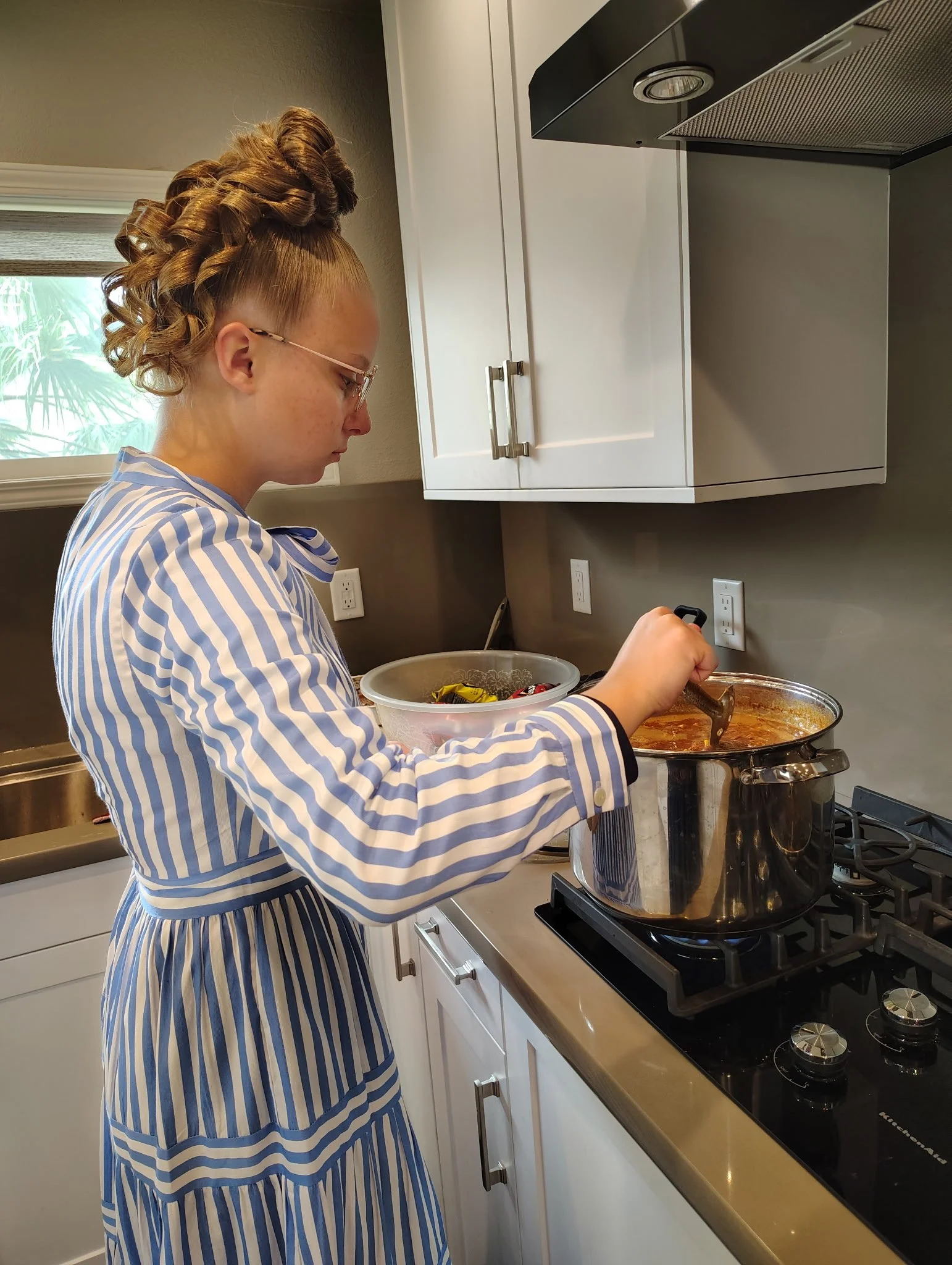 A young woman with blonde hair styled in curls and wearing a blue and white striped dress is cooking on a stovetop in a kitchen.