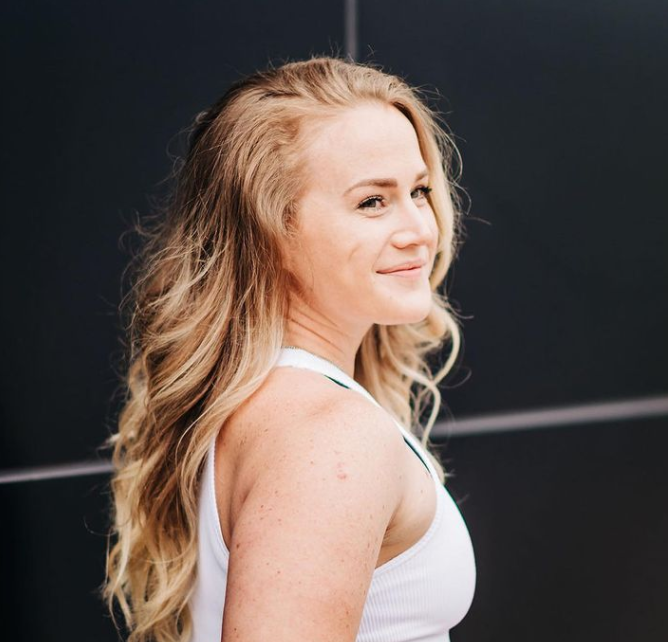 A woman with long wavy blonde hair smiling at the camera, wearing a white sleeveless top, standing outdoors against a dark background.