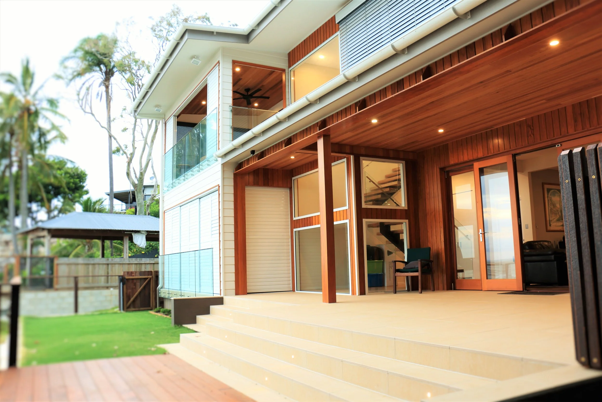 Exterior view of a modern two-story house with wooden accents, glass balcony, and sliding glass doors, with steps leading to a patio and a backyard with a gazebo and green lawn.
