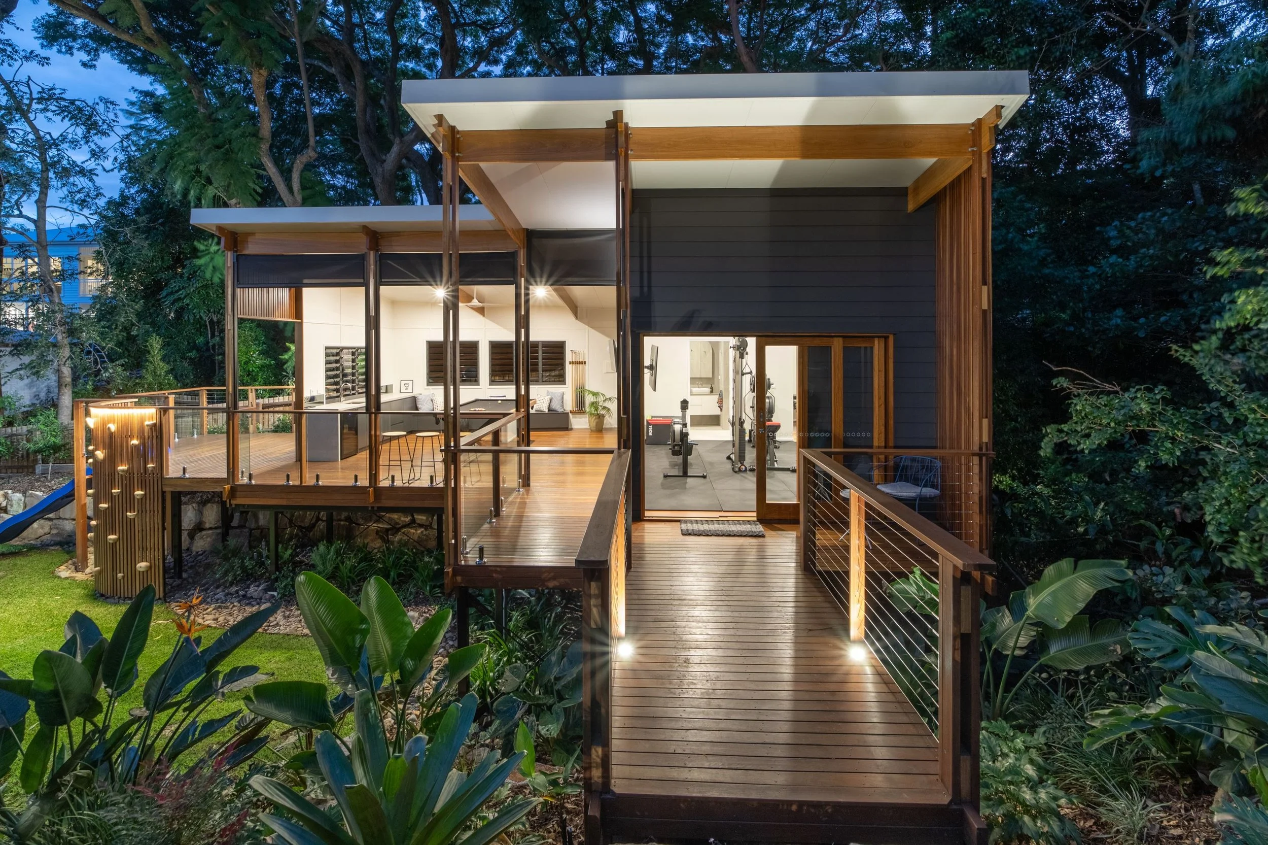 A modern two-story house with a wooden deck, large glass windows, and a gym inside, surrounded by lush greenery at dusk.