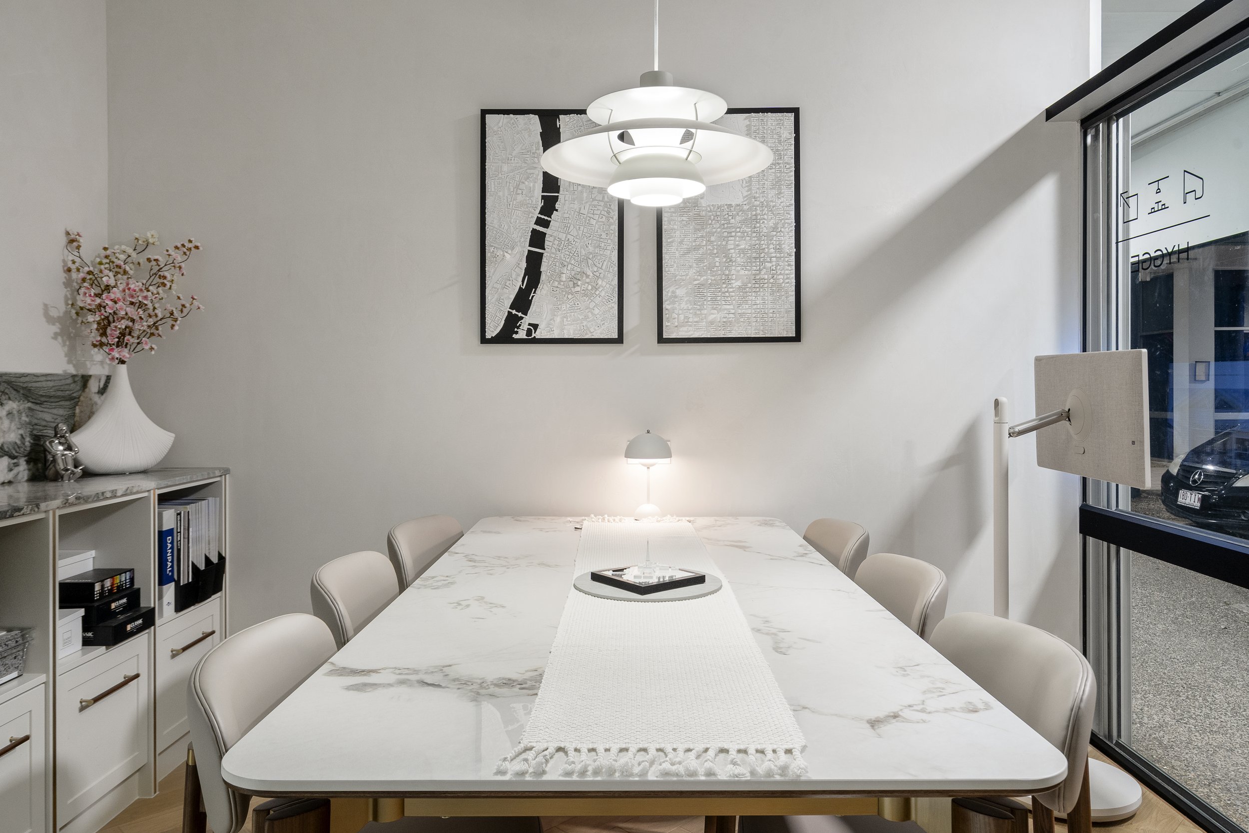A modern dining room with a white marble table, surrounded by white chairs, a hanging ceiling light, and framed maps on the wall. There is a window with a view outside.