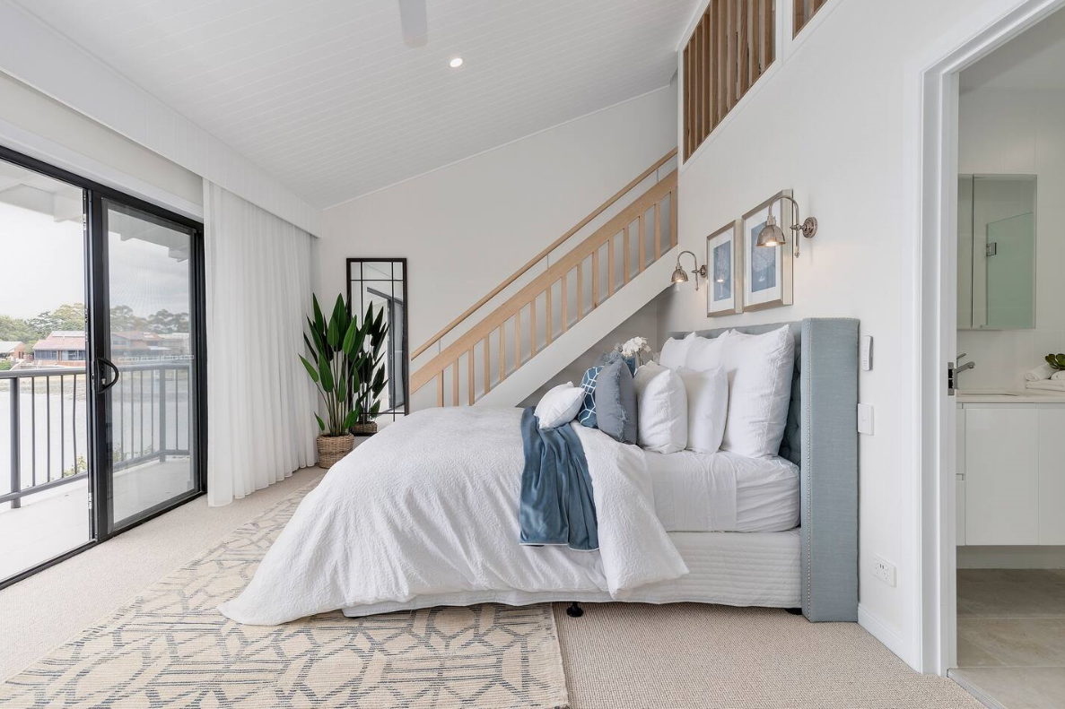 Bright bedroom with a bed, multiple pillows, and a blue throw blanket, next to a sliding glass door with white curtains, a potted plant, and a staircase