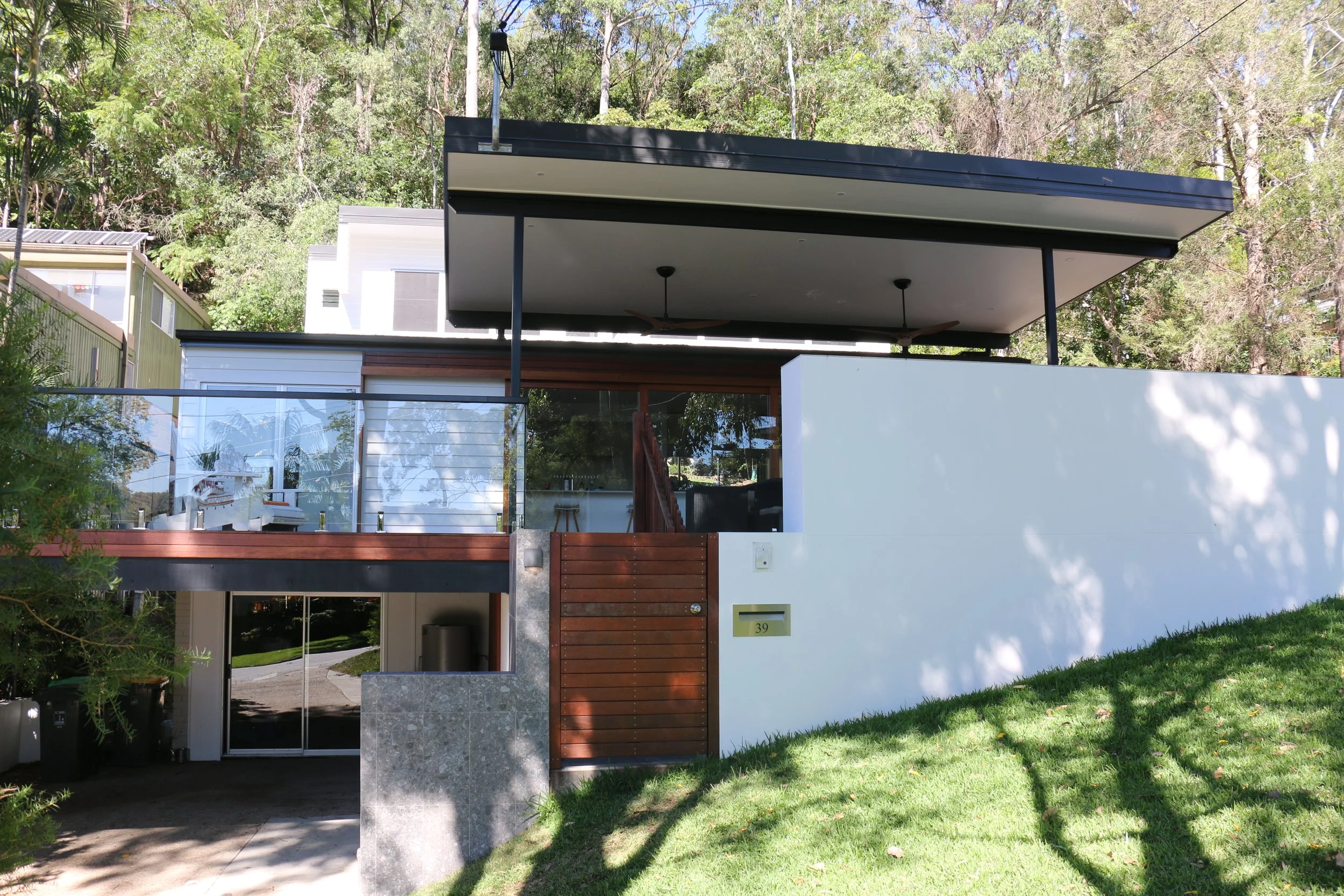 Modern two-story house with white exterior walls, wooden accents, a glass balcony, and a flat roof, surrounded by greenery.