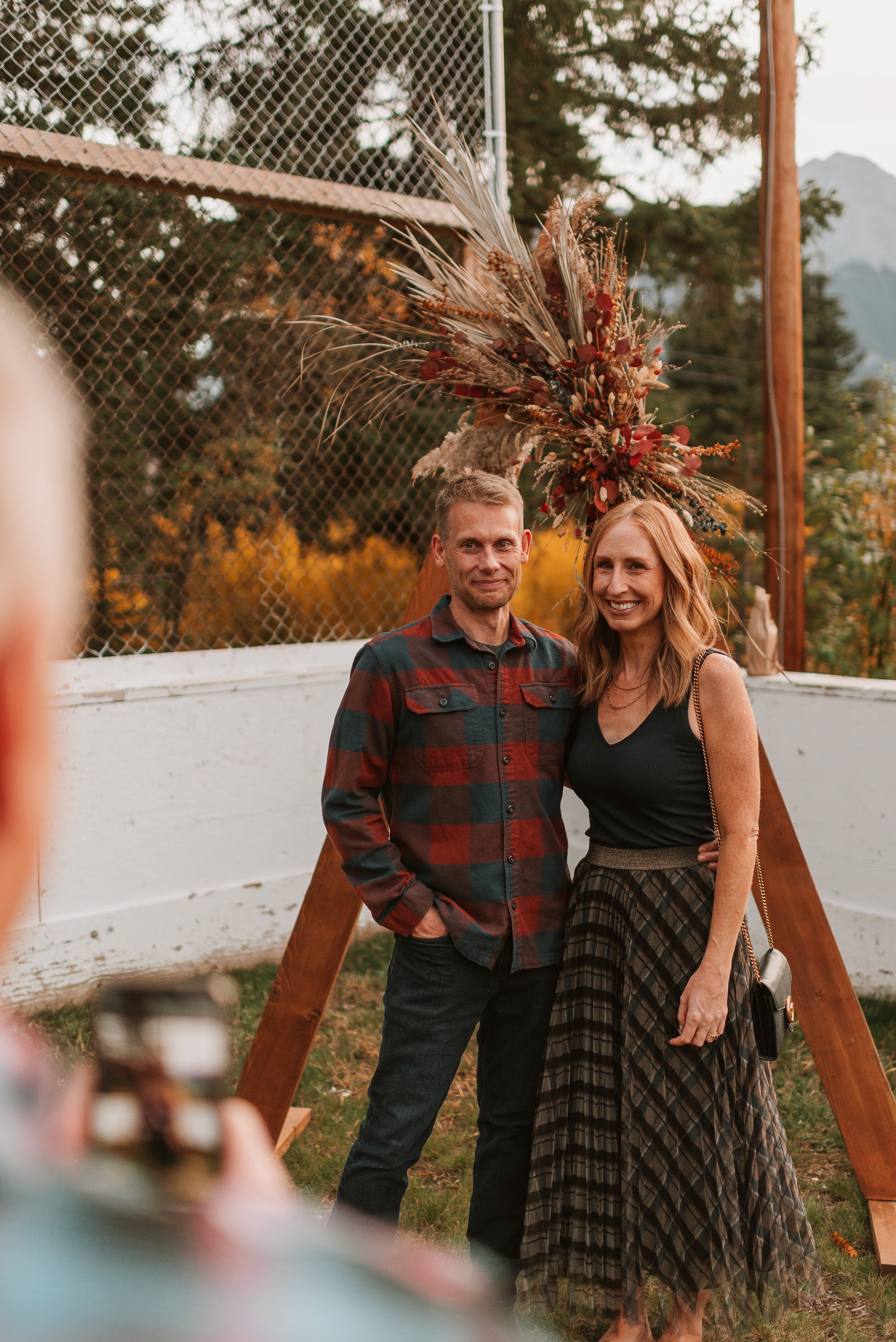A man and woman standing outdoors in front of a large floral arrangement, smiling for a photo.