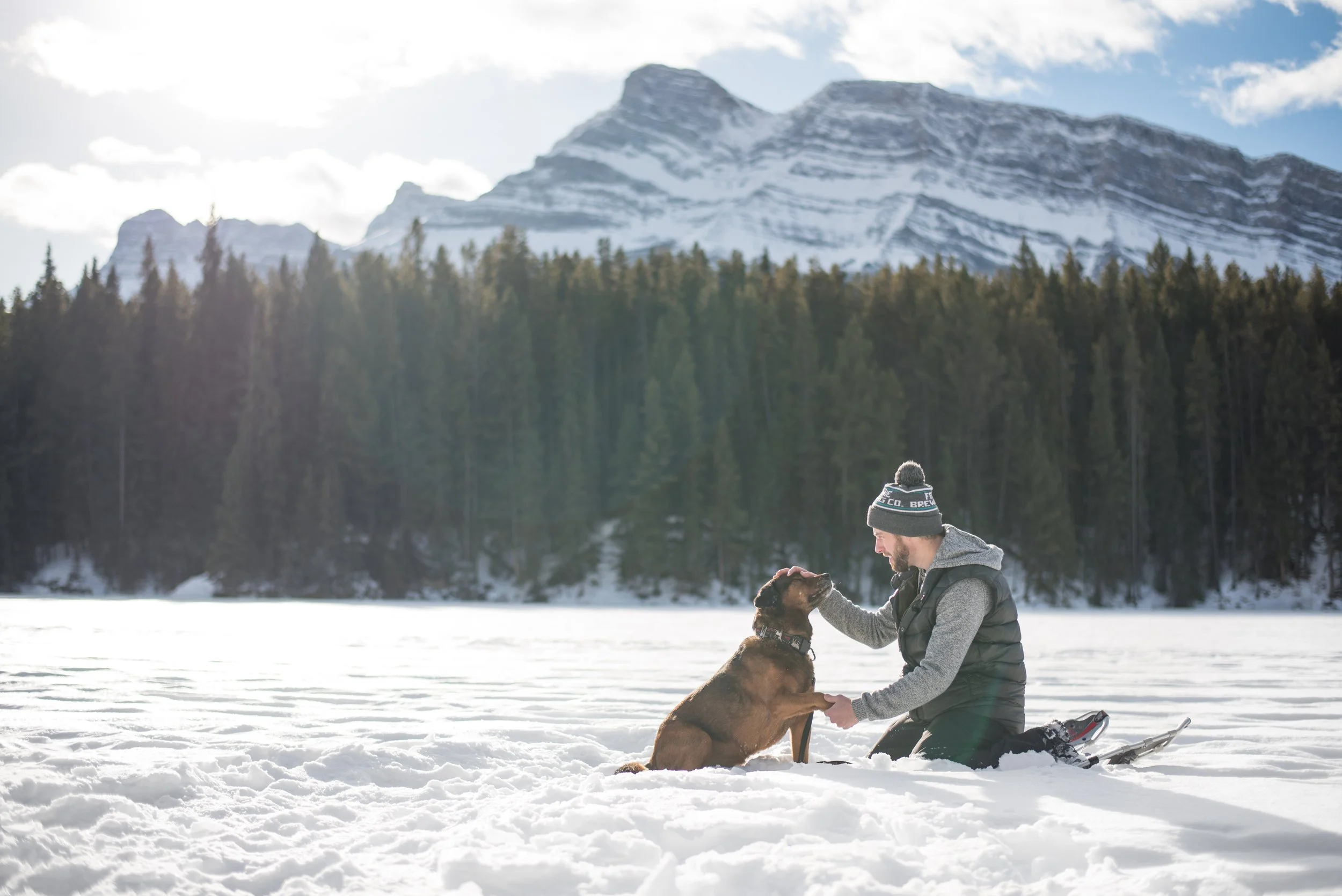 A man in winter clothes, kneeling on snow, petting a brown dog near a frozen lake with mountain and forest background.