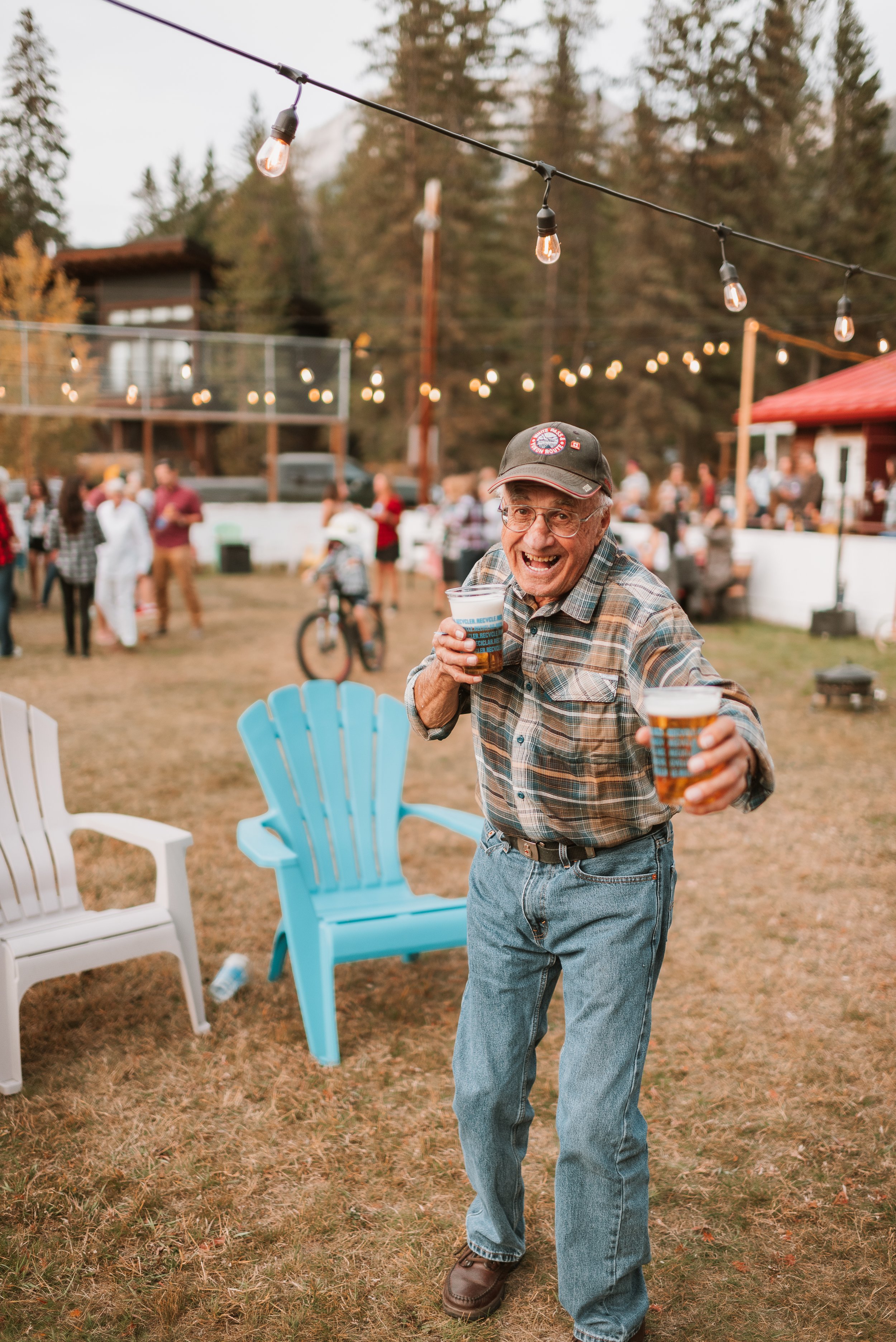An elderly man with glasses, wearing a plaid shirt, jeans, and a cap, smiling and holding two plastic cups of beer at an outdoor gathering with string lights, other people, and trees in the background.