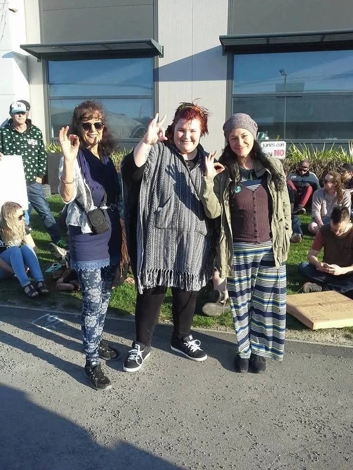 Three women standing outdoors in front of a building, smiling and making hand gestures, with a group of people sitting behind them on the grass.