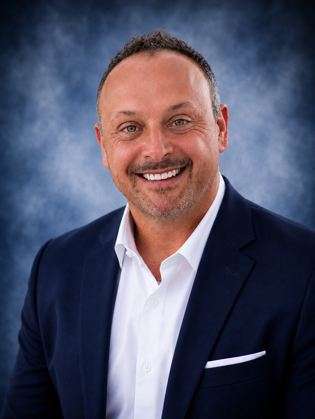 A man with short brown hair and blue eyes is smiling slightly at the camera. He is wearing a navy blue athletic jacket with a white logo that reads "Travel Desk Inc GOING EVERYWHERE NATIONWIDE" on the chest. The background is a blurred dark and gray textured backdrop.