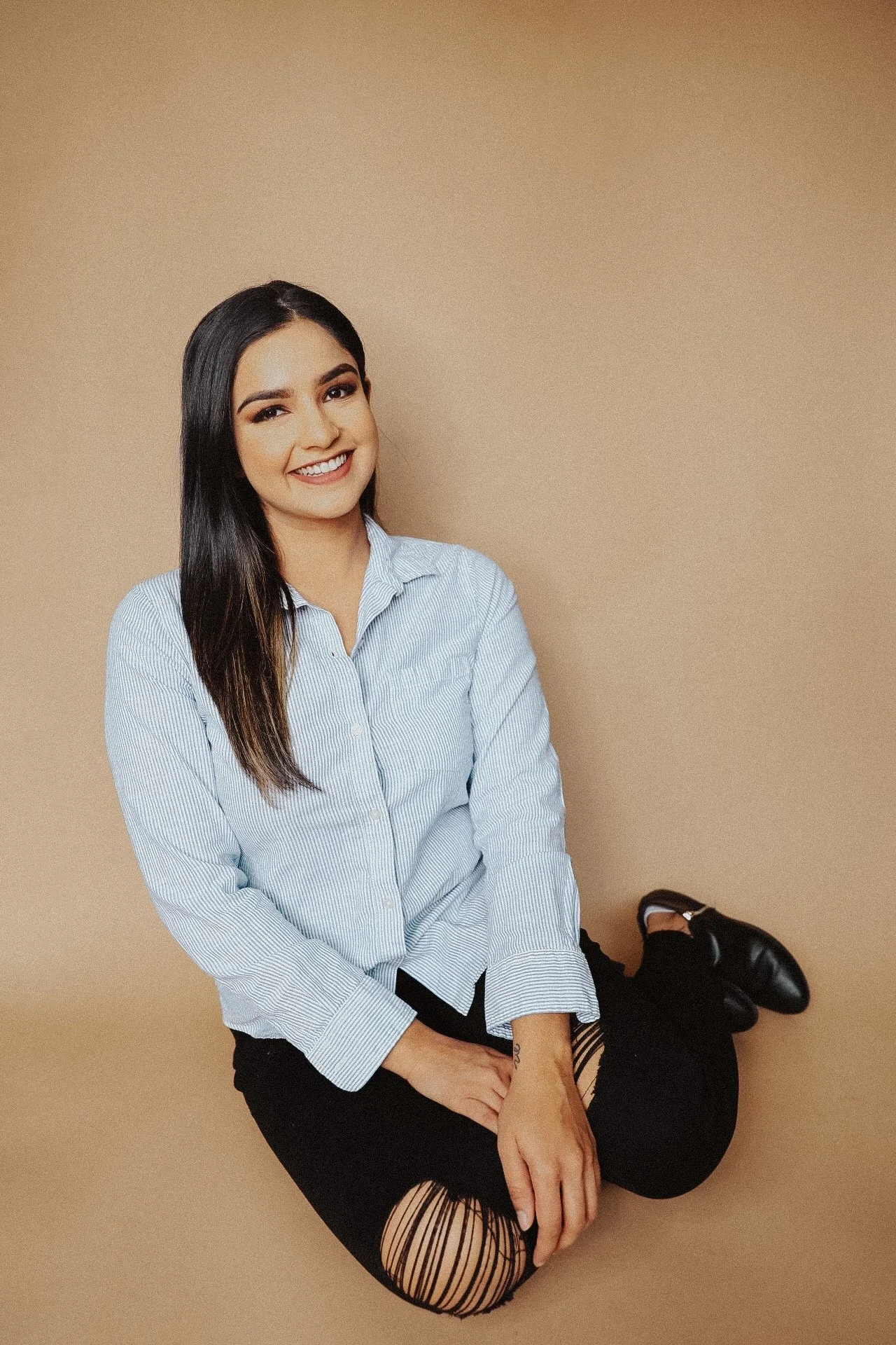 A young woman with long dark hair wearing a light blue striped button-up shirt, black ripped jeans, and black shoes, sitting on the floor and smiling at the camera against a beige background.