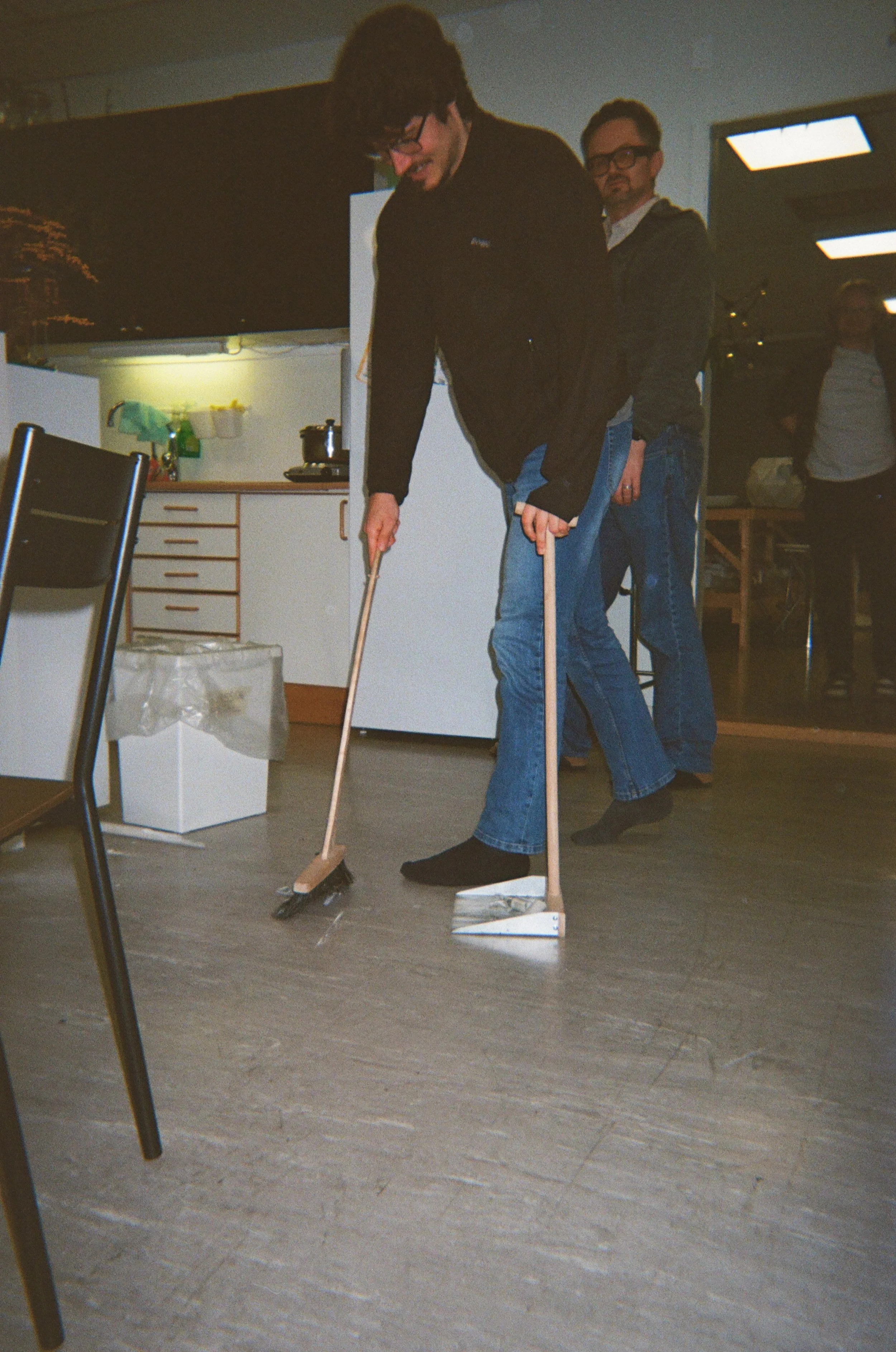 To personer med blå jeans og svarte sko står på gulvet med mop og feiekost i hånden.