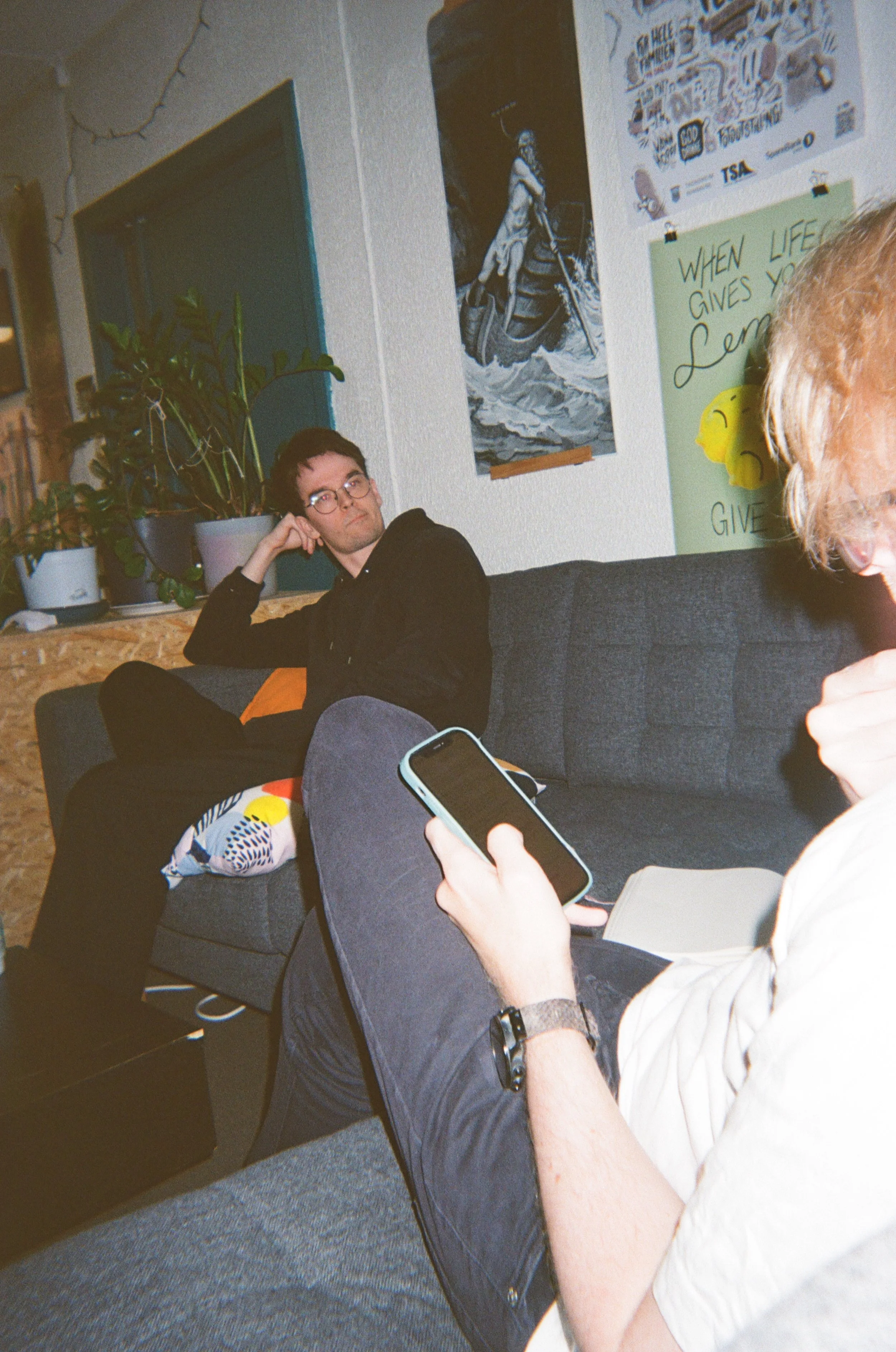 To personer som sitter på en sofa i et rom, hvorav en person bruker telefonen sin og den andre ser på. Rommet har plakater på veggene.