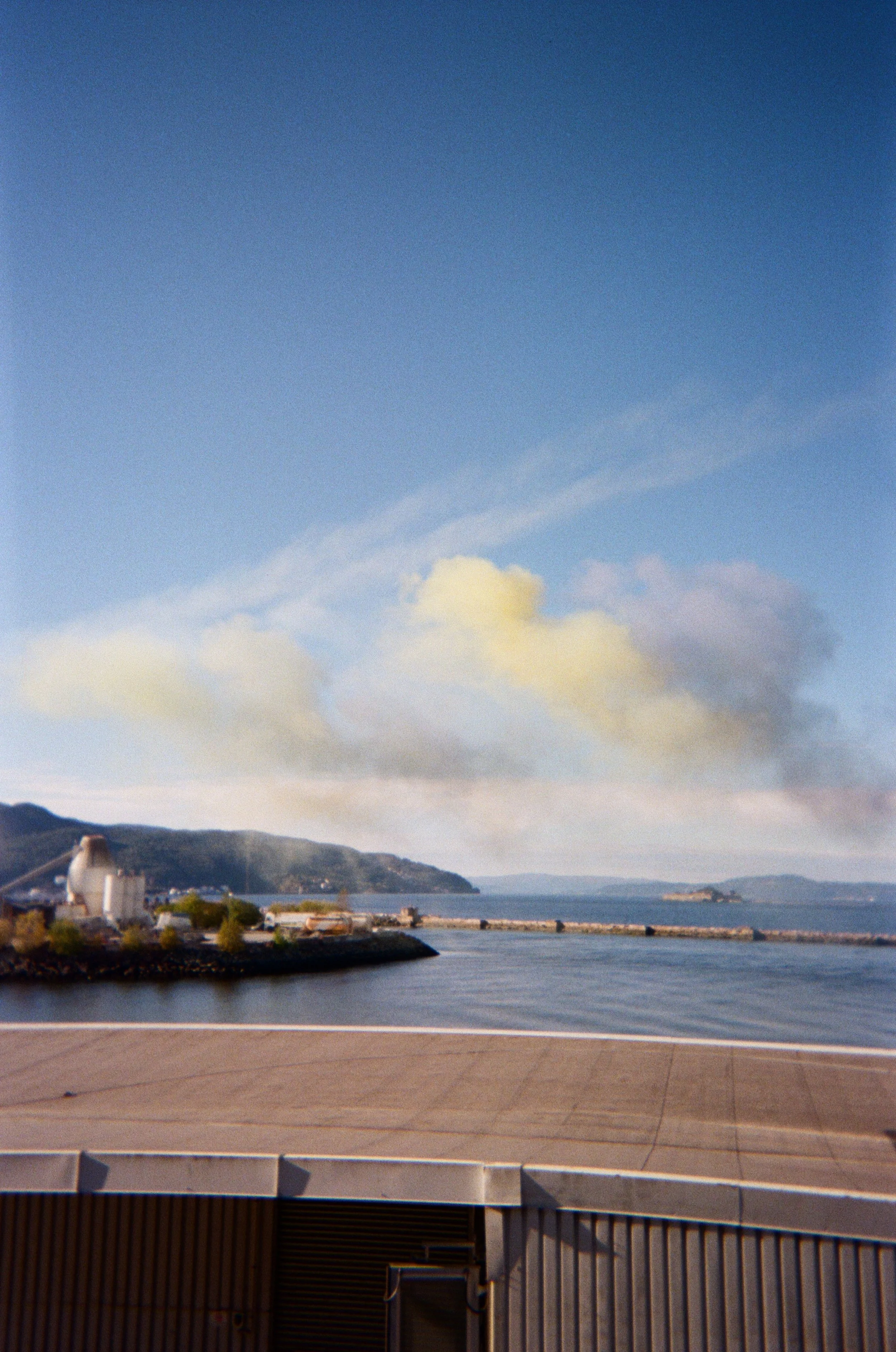 Utsikt over havn med bygninger og en samling av bygninger i fjellet i bakgrunnen. Klar blå himmel med noen skyer.