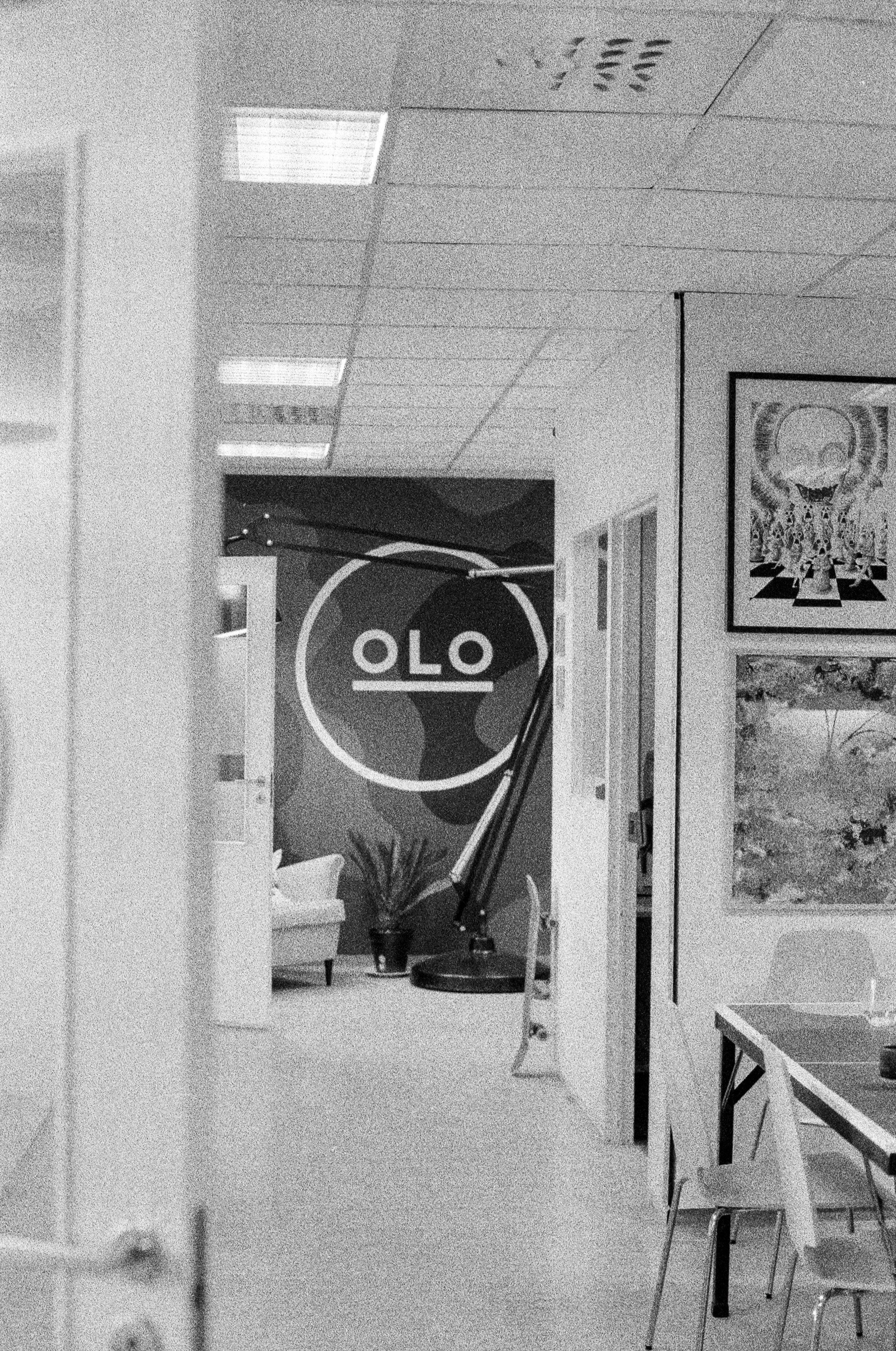Interiørbilde av et kontormiljø med en vegg med OLO-logo, planter, en stol, og kunst på veggene, i svart-hvitt.