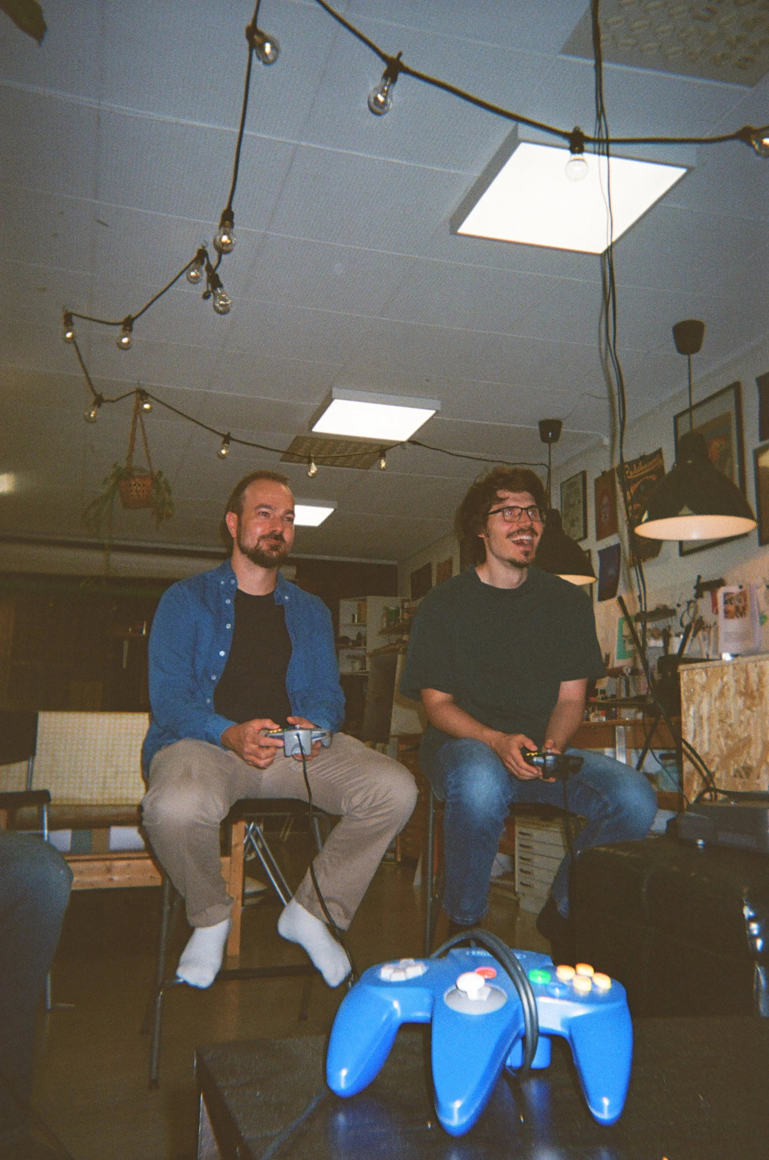 To menn spiller videospill sammen i et rom, med en spillkontroller i hendene, mens de sitter på stoler. Det er en lyskrans hengt opp i taket og en blå spillkontroller foran dem.