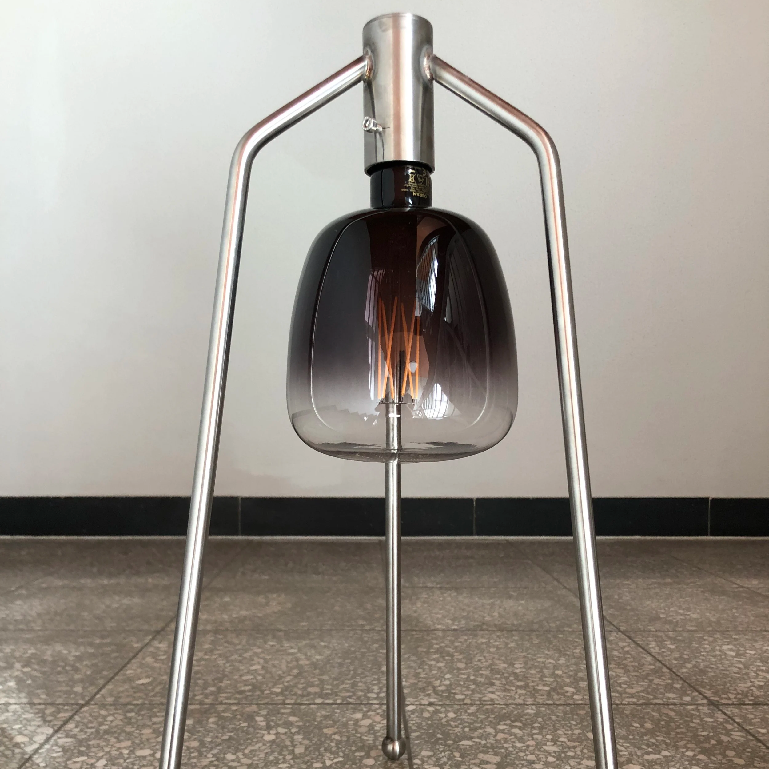 A distinctive pendant light fixture with a black glass shade and woven filament bulb, supported by a metallic frame, set against a neutral background, captured from a low angle.