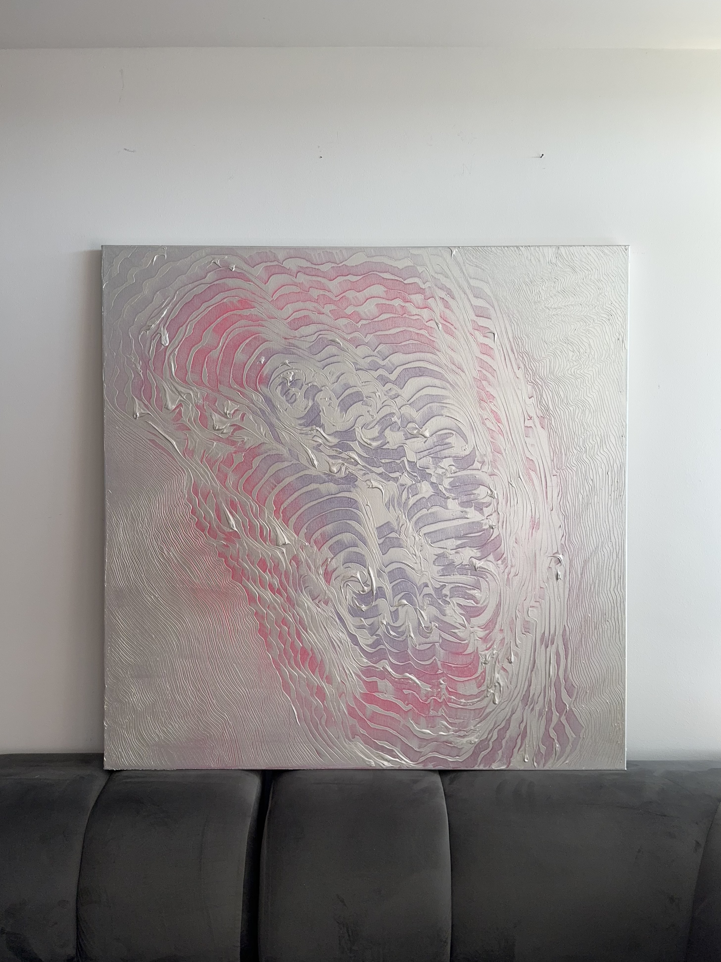 Abstract painting with swirling patterns in pink, purple, and silver hues, displayed on a wall above a black sofa.