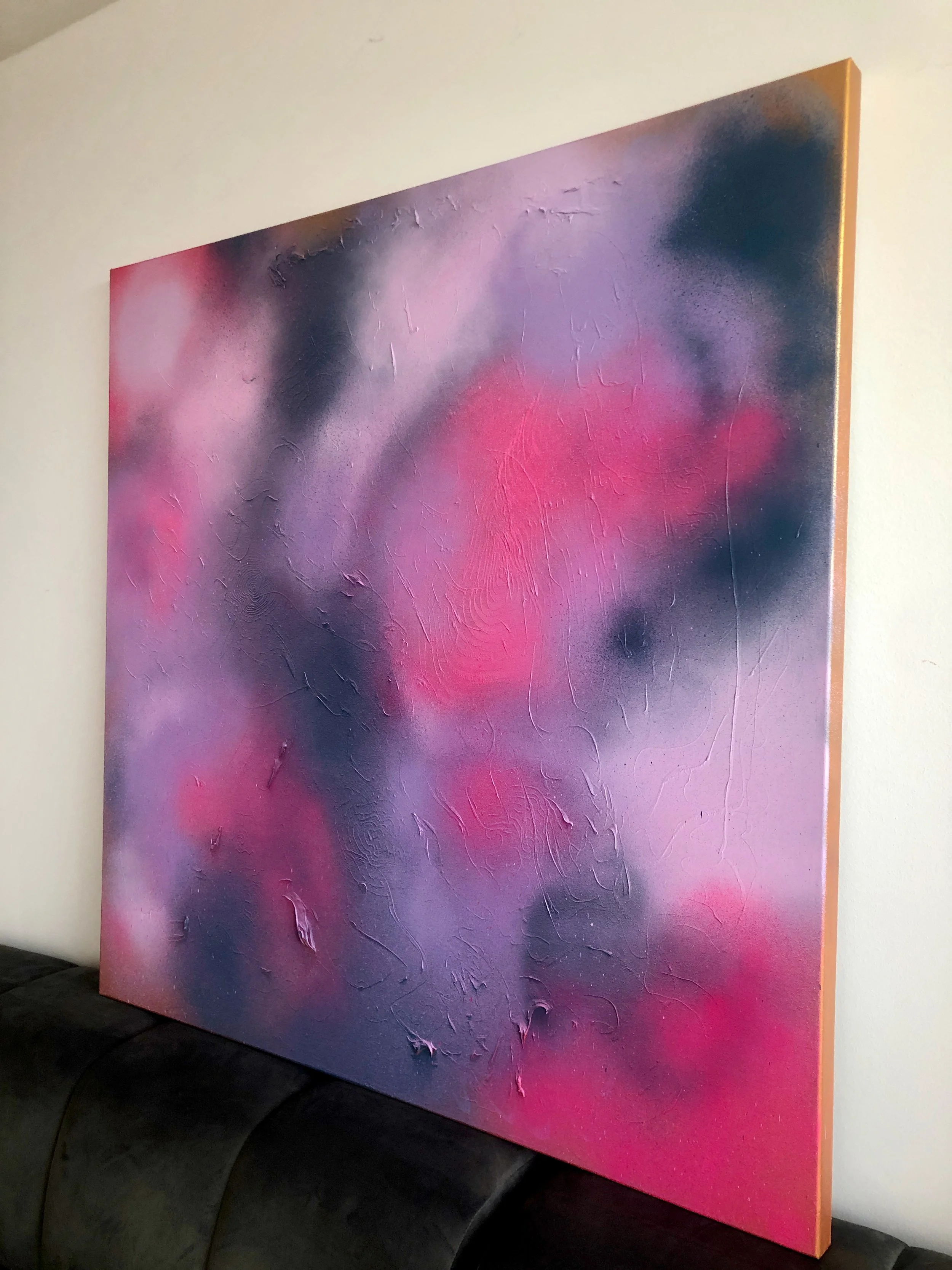 Close-up of an abstract, textured painting with pink, purple, black, and orange colors on a canvas leaning against a white wall.