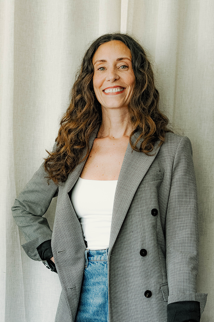 Woman with long curly hair in a black buttoned dress smiling in front of a white brick wall.