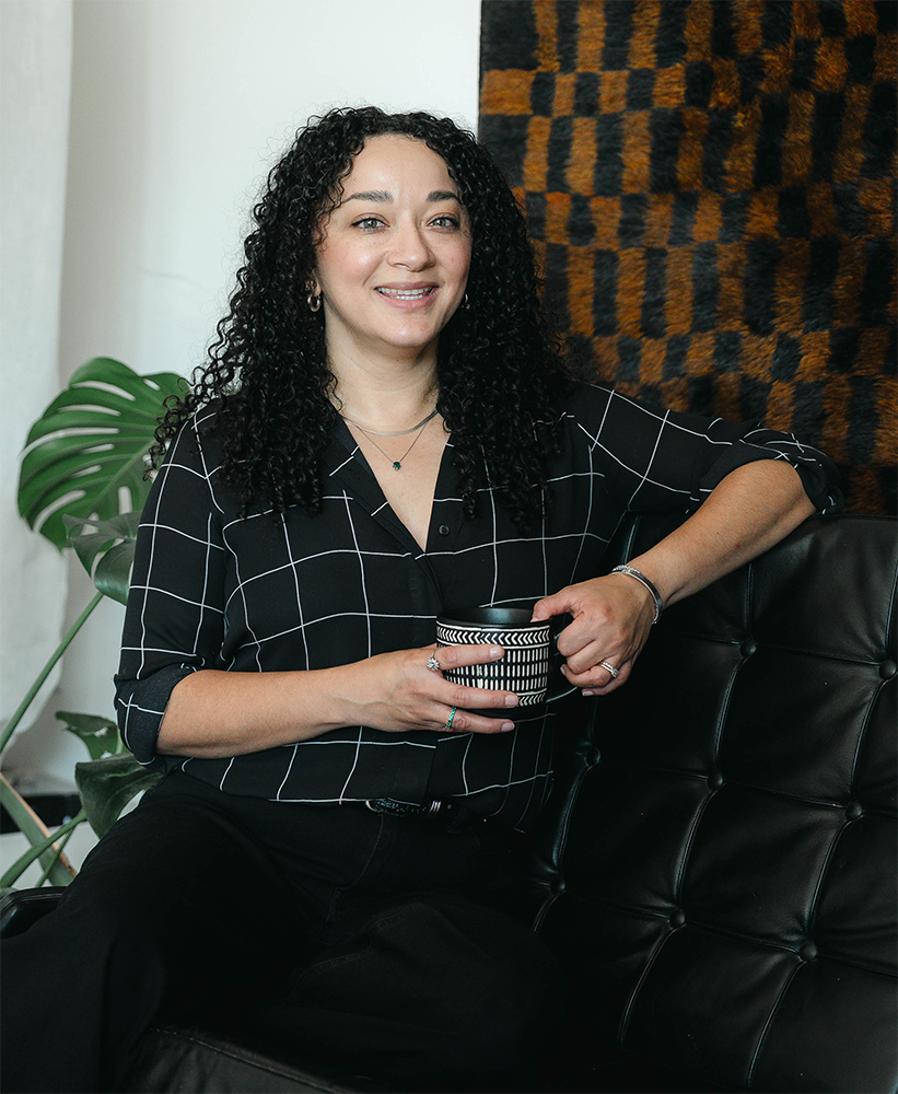 Person with curly hair wearing a black top posing against a white brick wall.