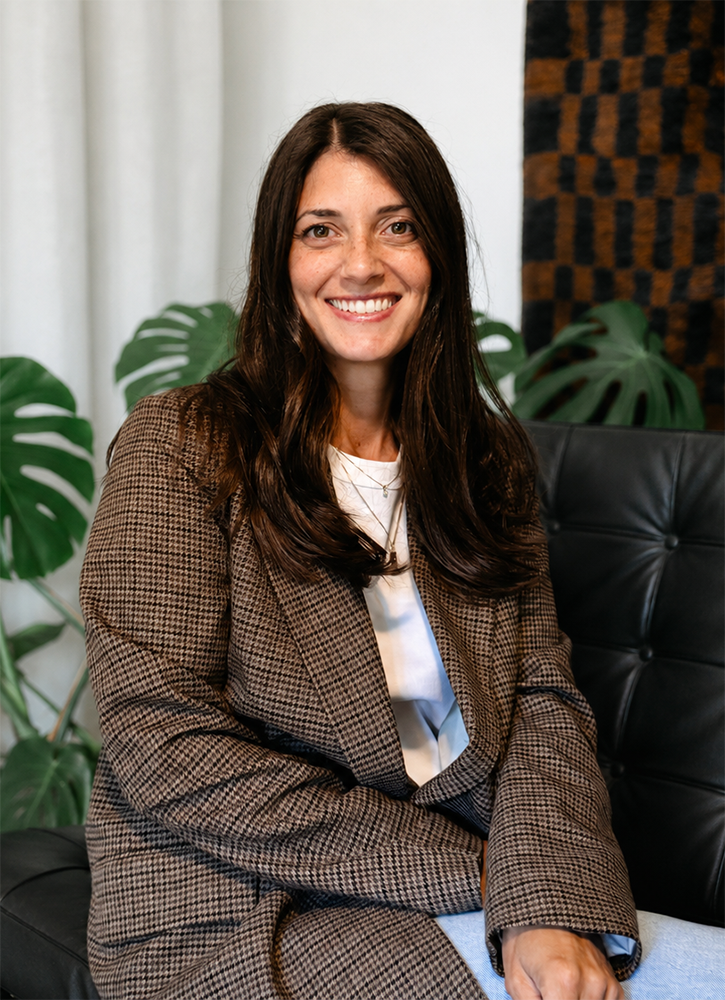 Smiling woman in black sweater with necklaces against white brick wall.